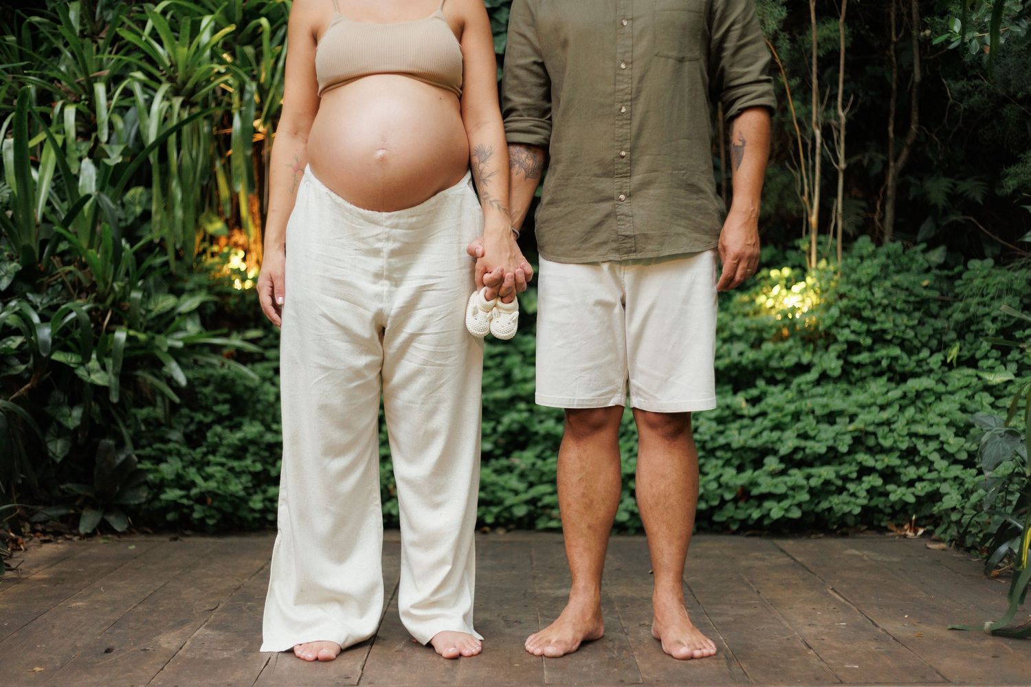 Pregnant couple standing barefoot holding tiny baby shoes in a green garden, torsos only visible.