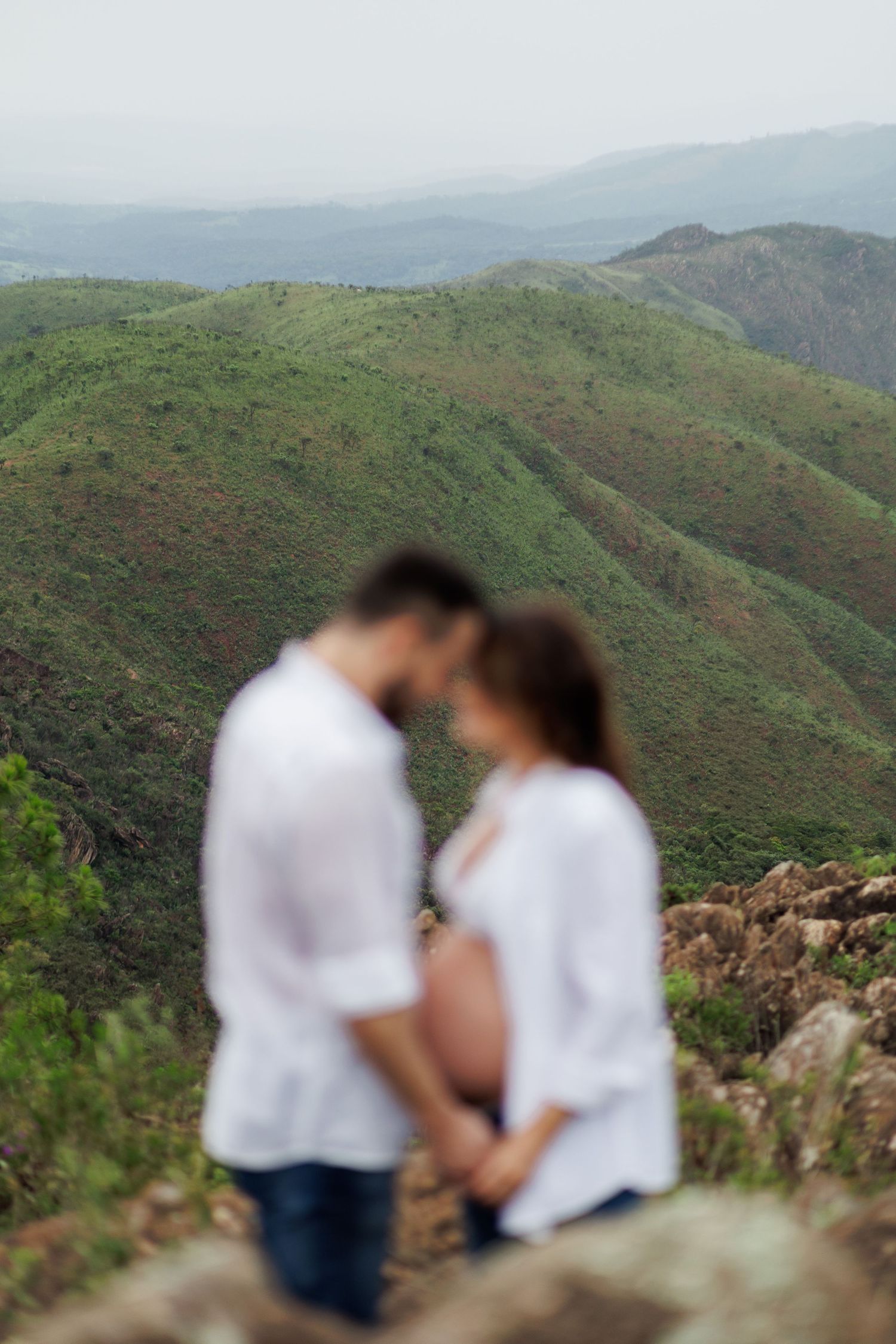 Blurred couple in white shirts sharing tender moment on mountain overlook with lush green hills backdrop.