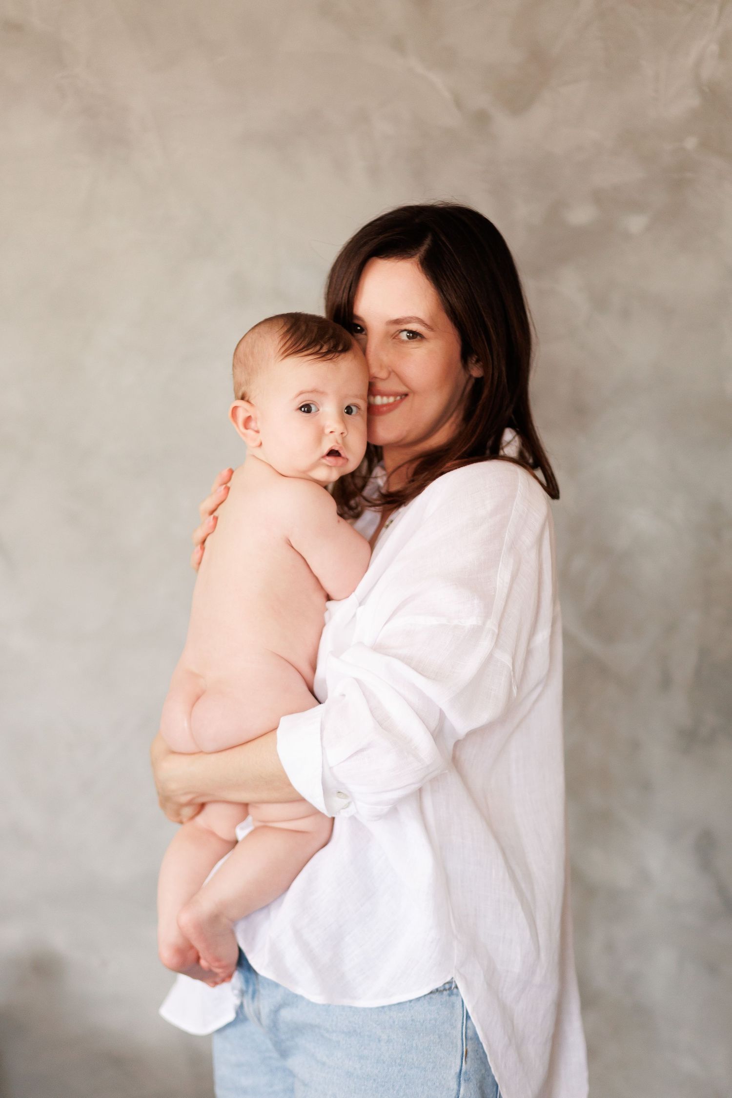 A smiling brunette mother in a white shirt holds her adorable baby against a gray textured wall.
