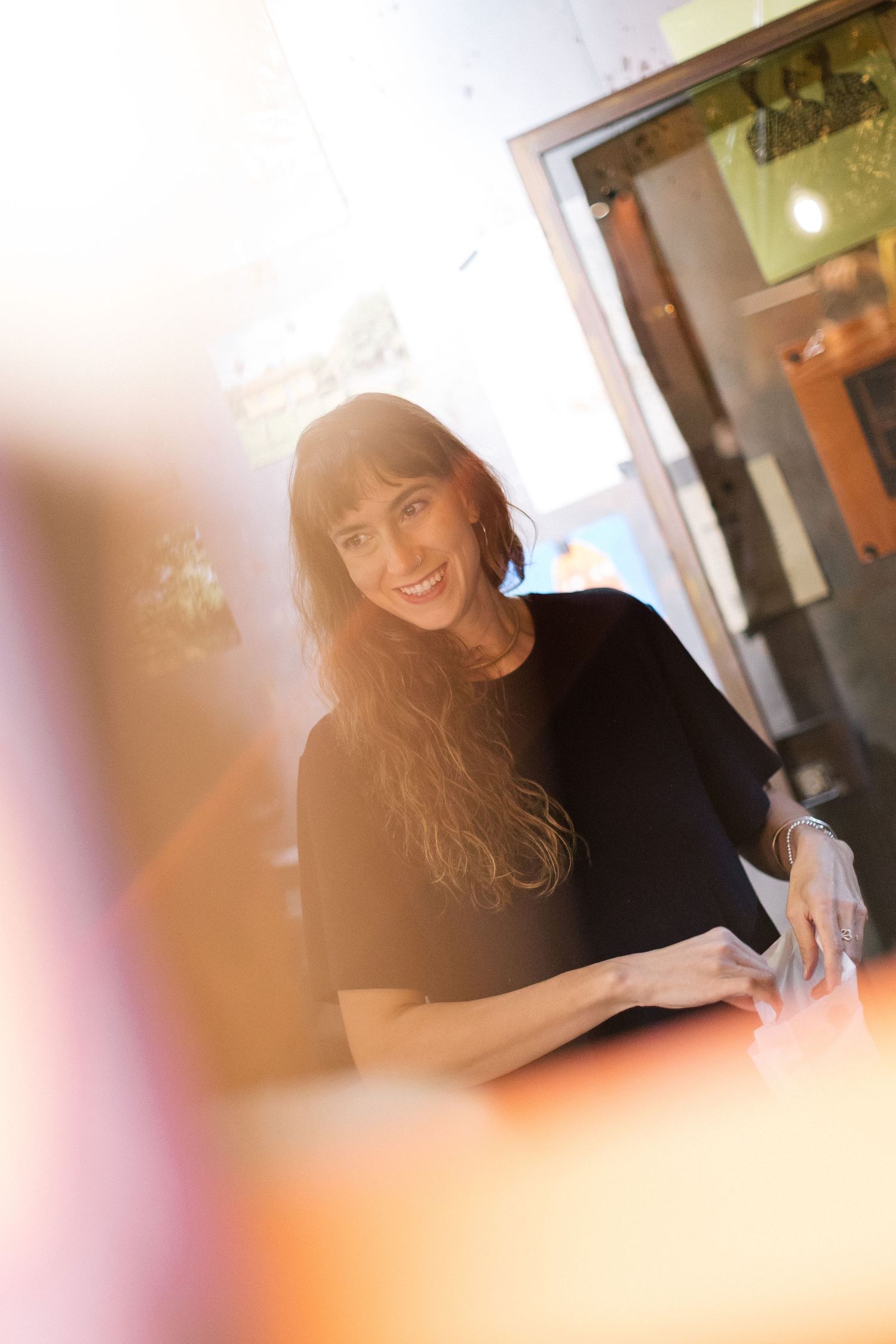 Smiling woman with long brown hair wearing a black top, leaning on a counter in a warmly lit indoor setting.
