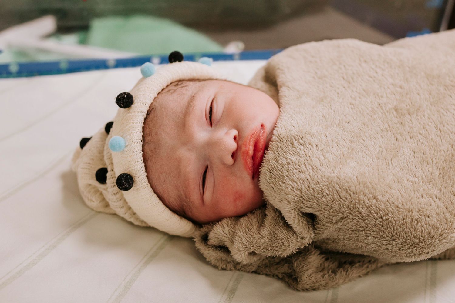 Peaceful newborn swaddled in beige blanket wearing a polka dot hat rests on hospital bed.