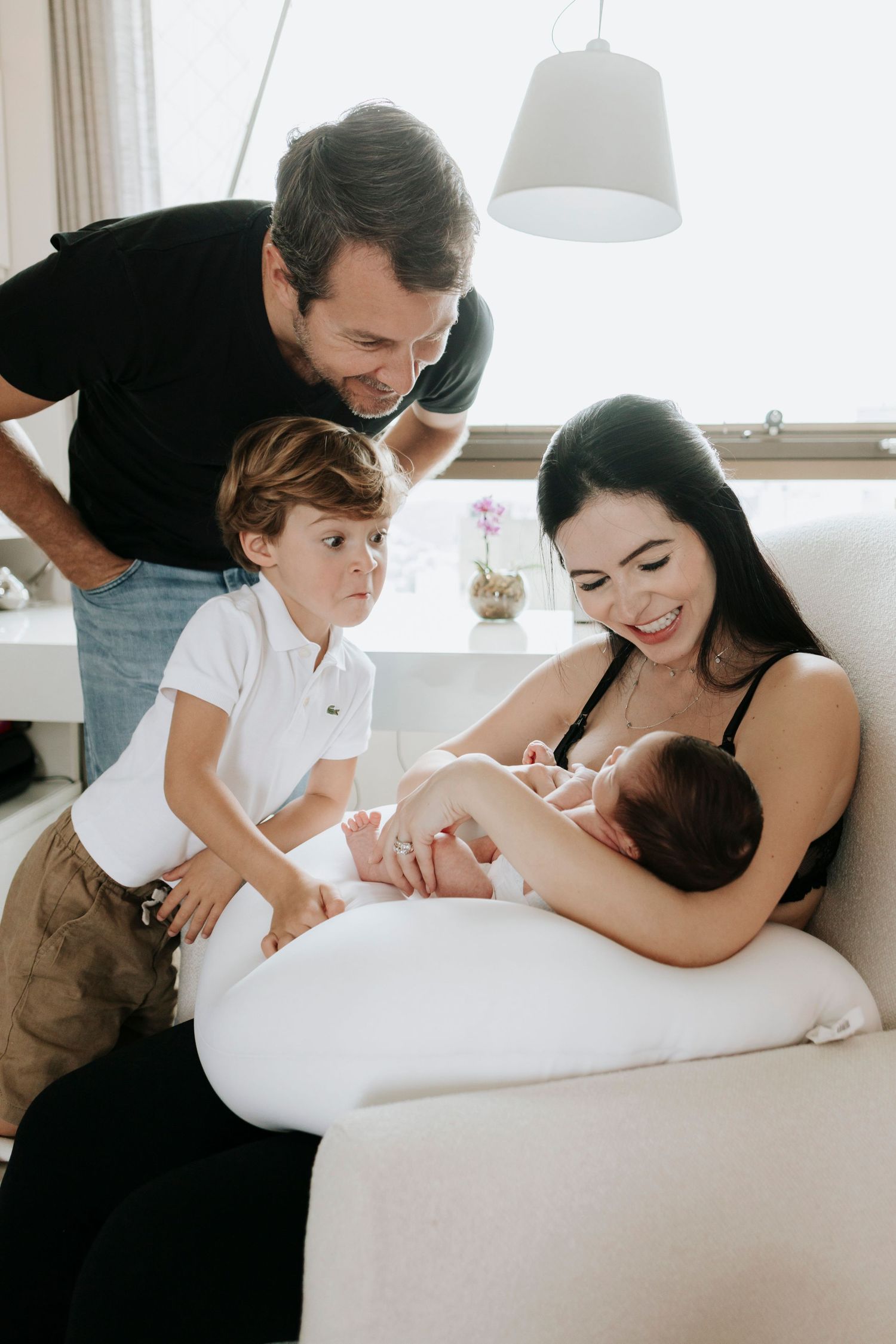 A family gathers around a nursing newborn baby on a white couch in a bright room.