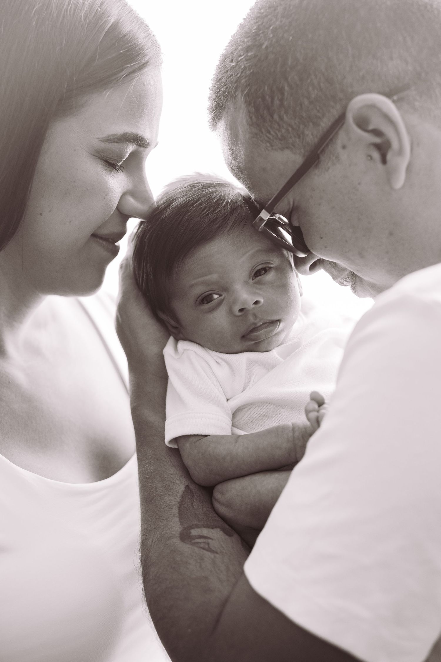 Black and white photo of parents lovingly cradling their newborn baby, touching foreheads together in tender family moment.