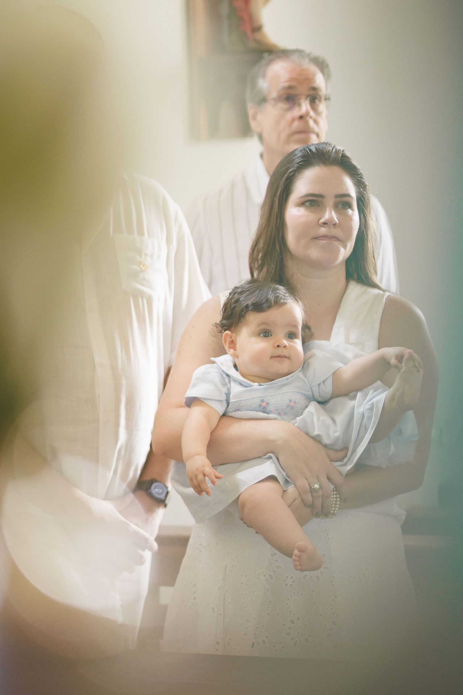 Woman in white dress holding baby at a baptism ceremony with an older man standing behind them.