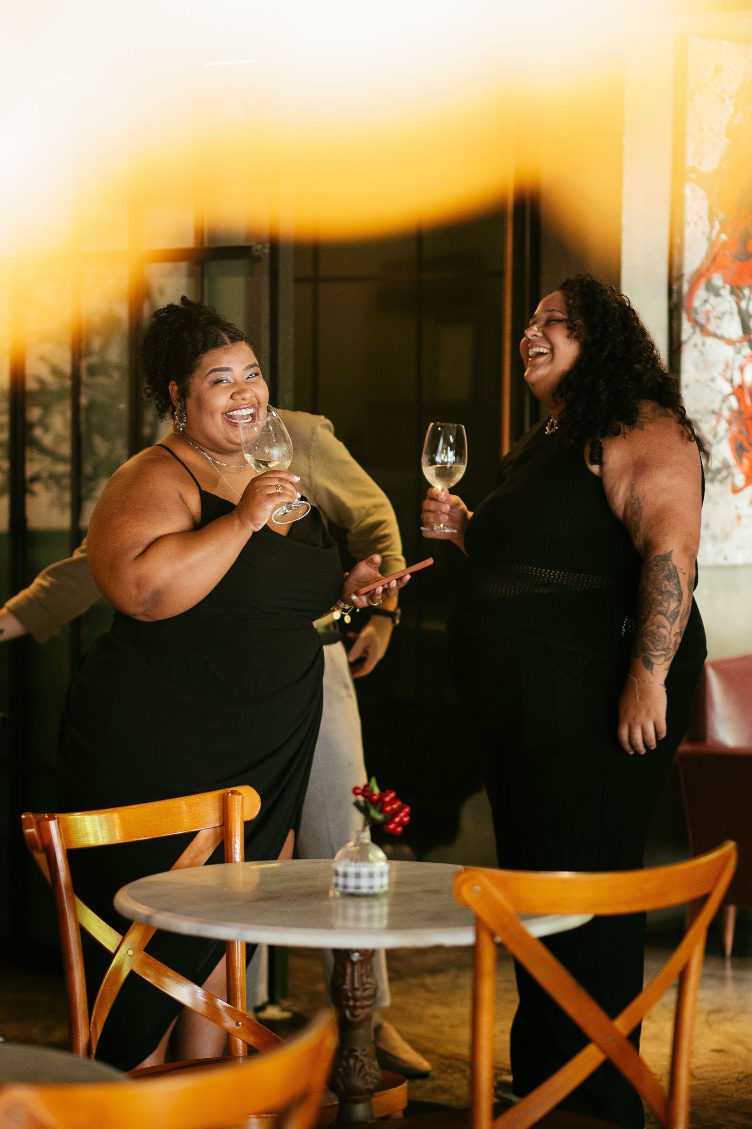 Two plus-size women laughing and holding wine glasses at a stylish indoor venue.