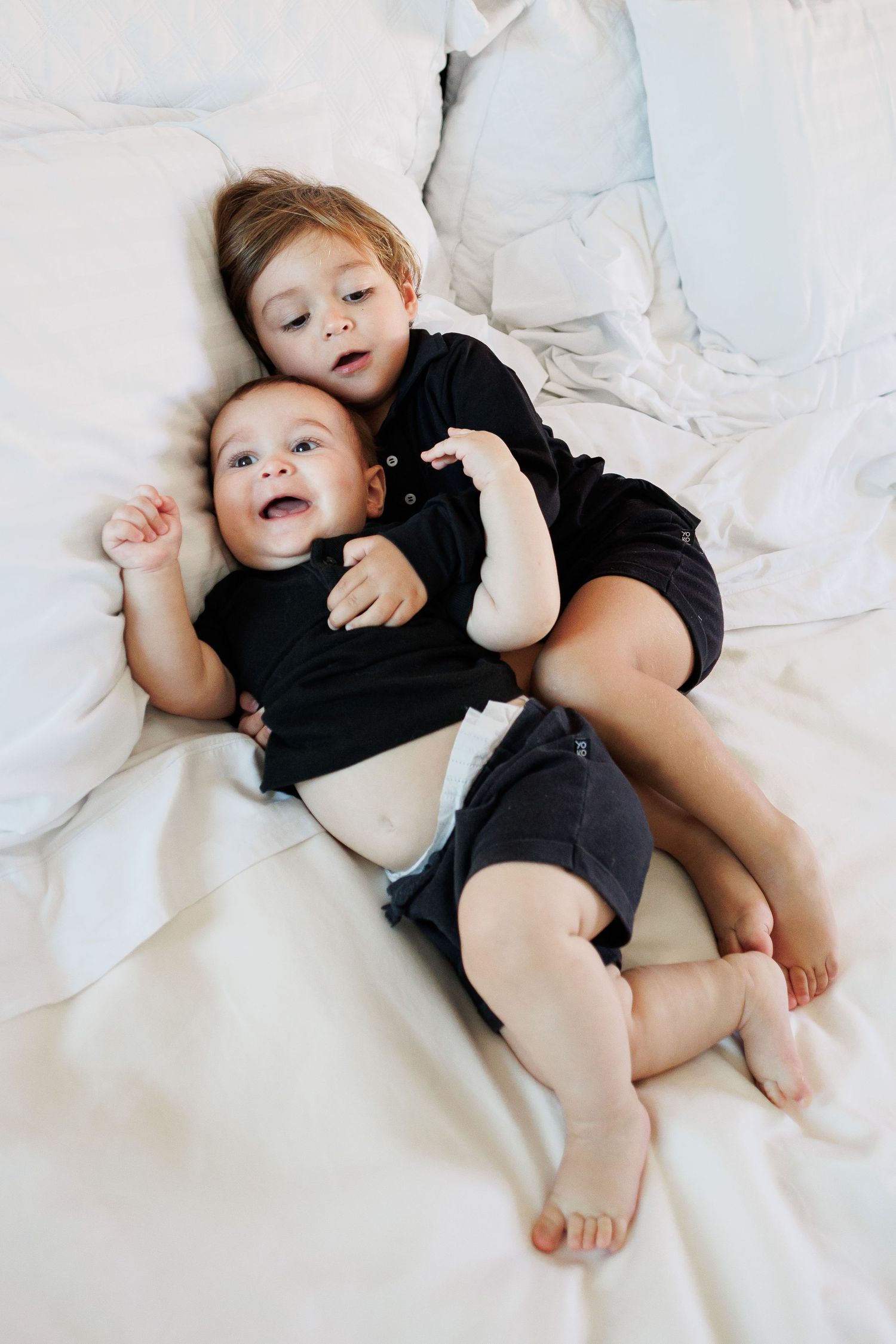 Two young siblings in matching black outfits cuddle together on a white bed, with the older child hugging the smiling baby.