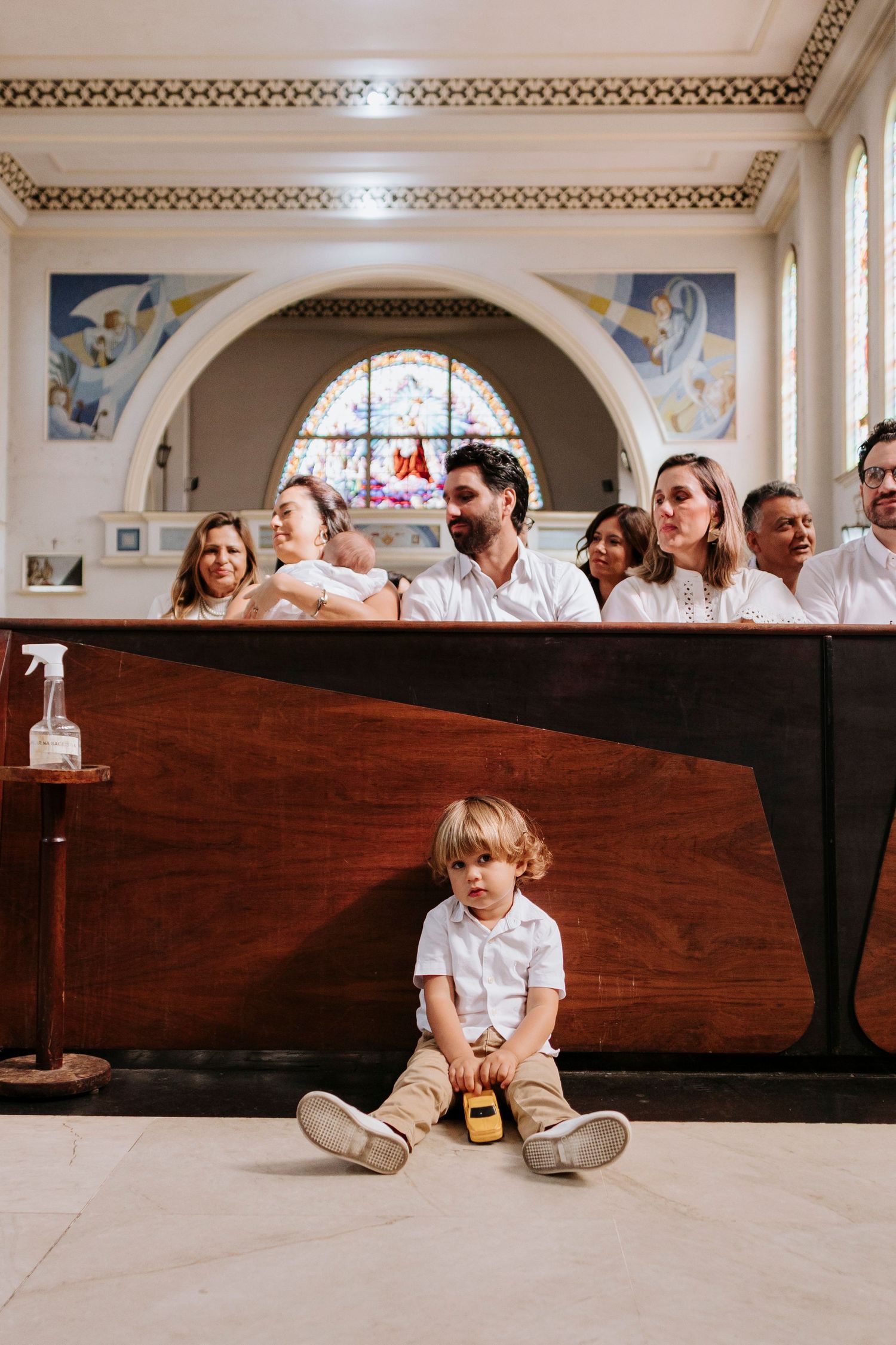 A small child in white sits alone on the floor in front of a wooden church pew while others sit behind during a service.