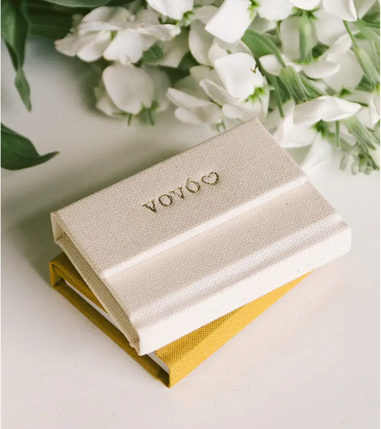 Elegant VOYÓ branded linen keepsake box in ivory with gold embossed logo, displayed with white flowers and yellow accent box.