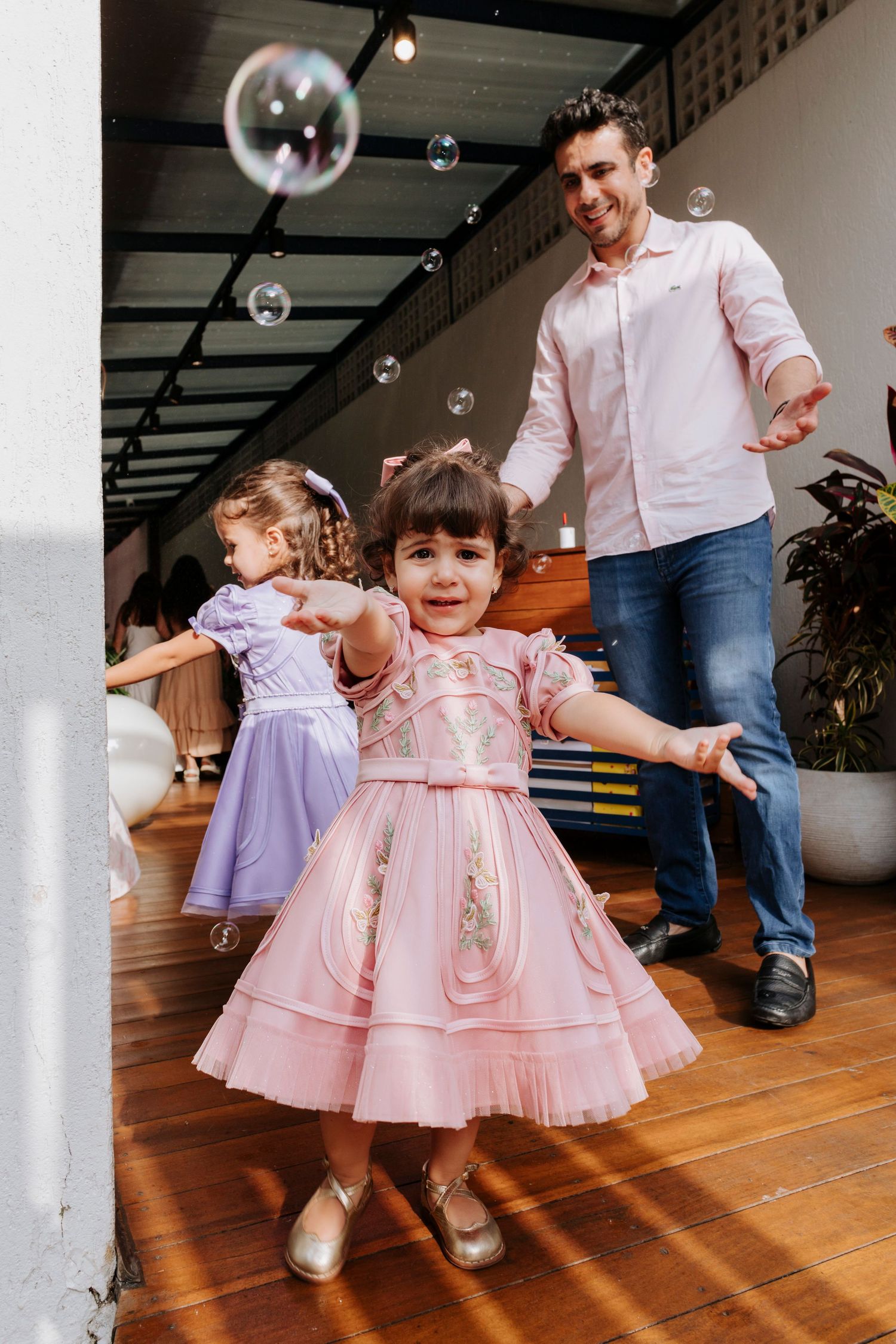 Children playing with bubbles in party dresses while dancing indoors.