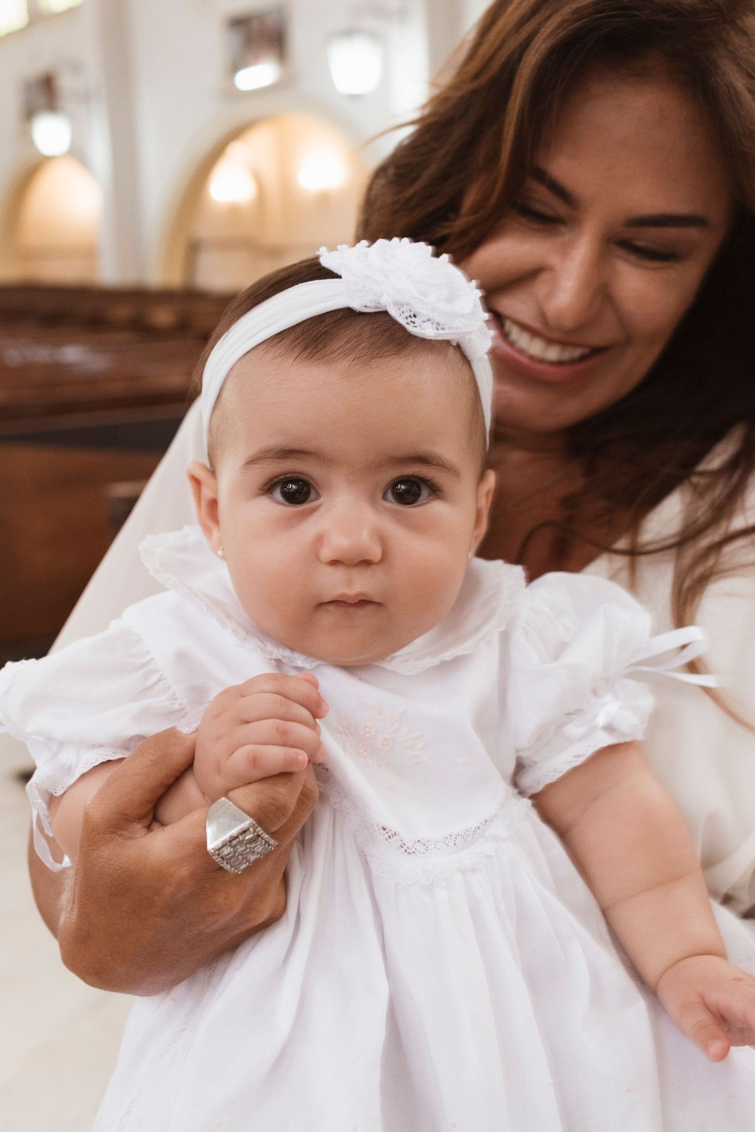 Baby in white christening gown and headband is held during baptism ceremony in church.