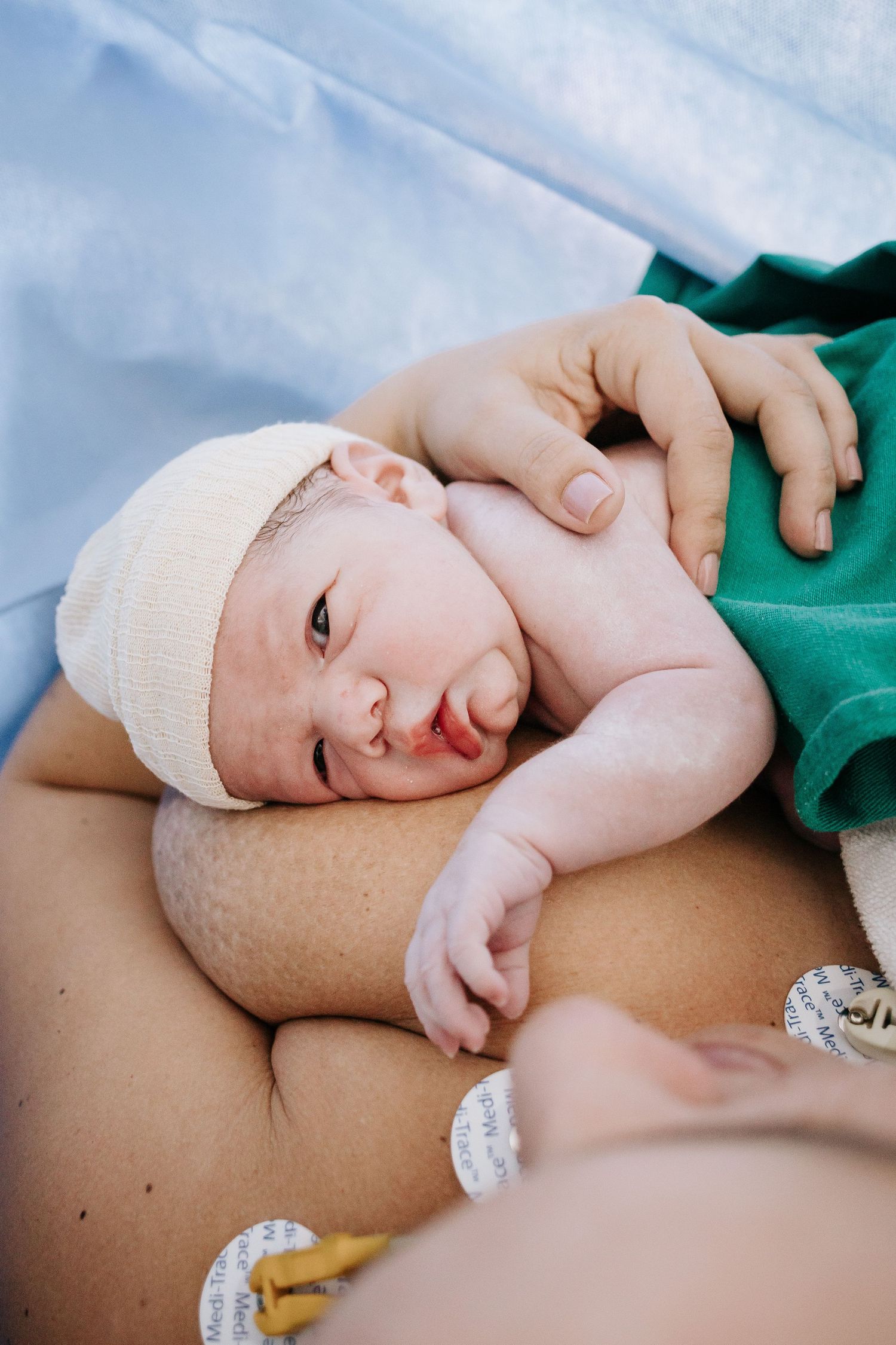 Newborn wearing white cap rests peacefully against soft blue hospital bedding.