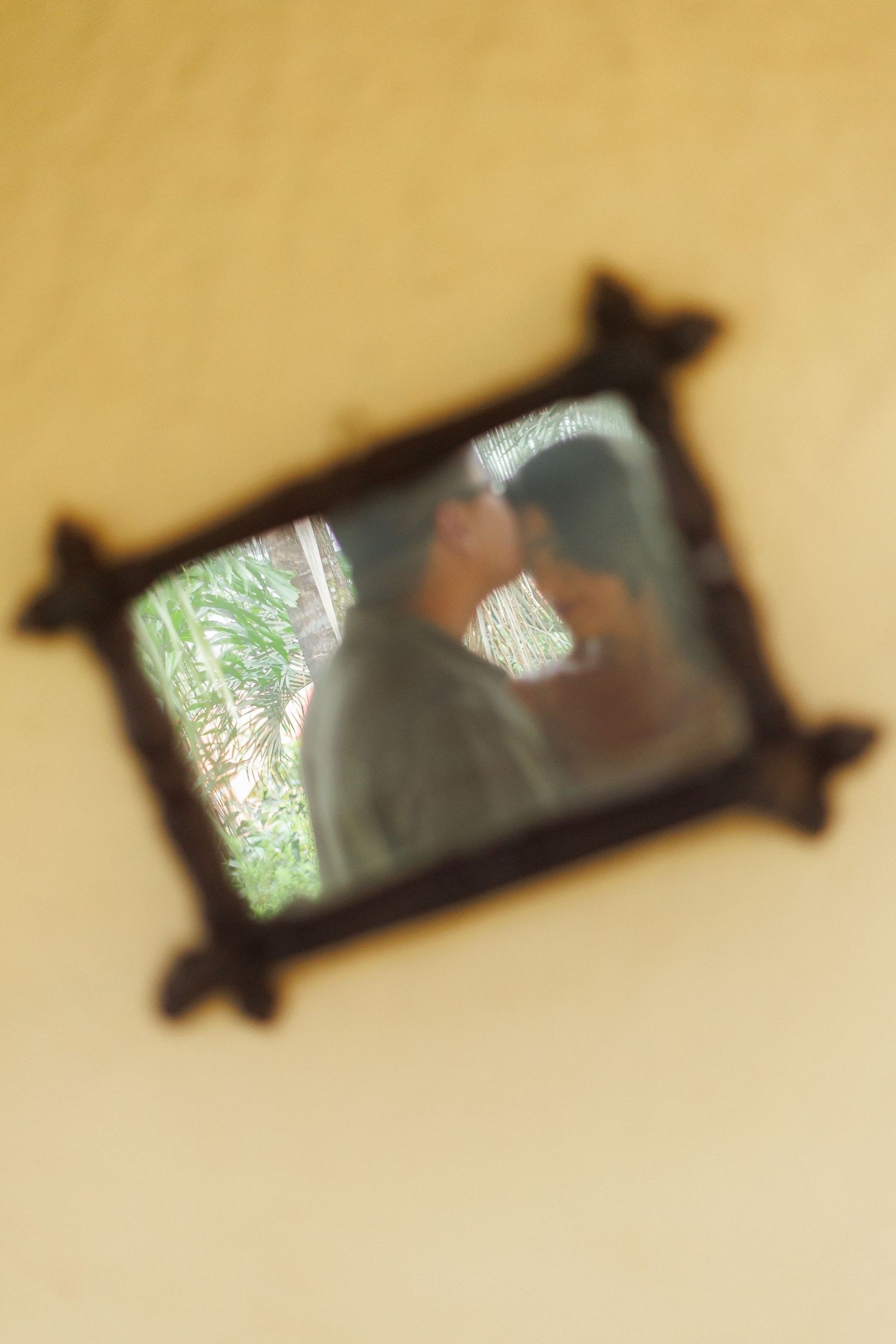 Blurry reflection of a couple kissing in an ornate dark wooden mirror hanging on a yellow wall.