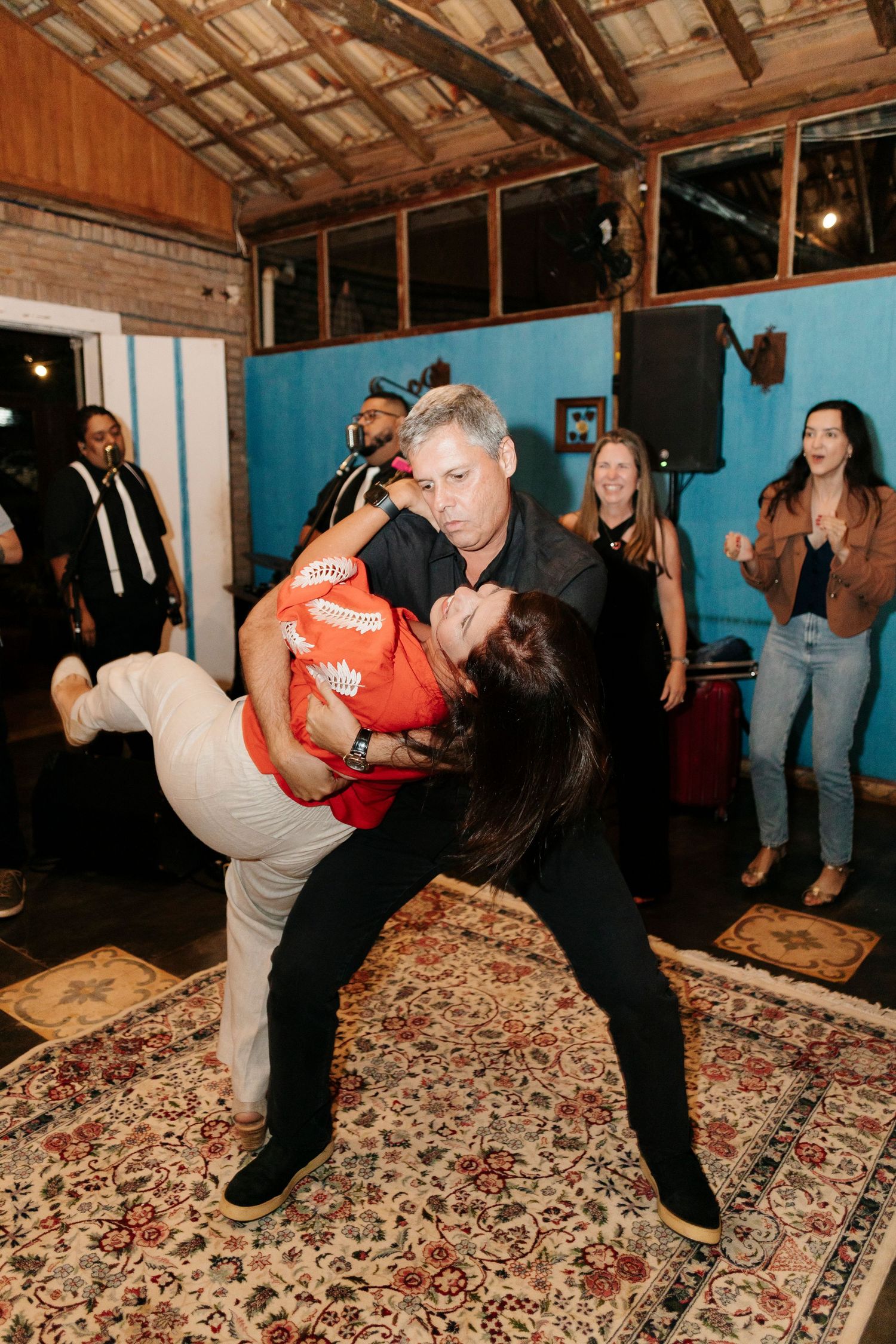 Dancers perform a dramatic dip move on a patterned rug at an indoor celebration.