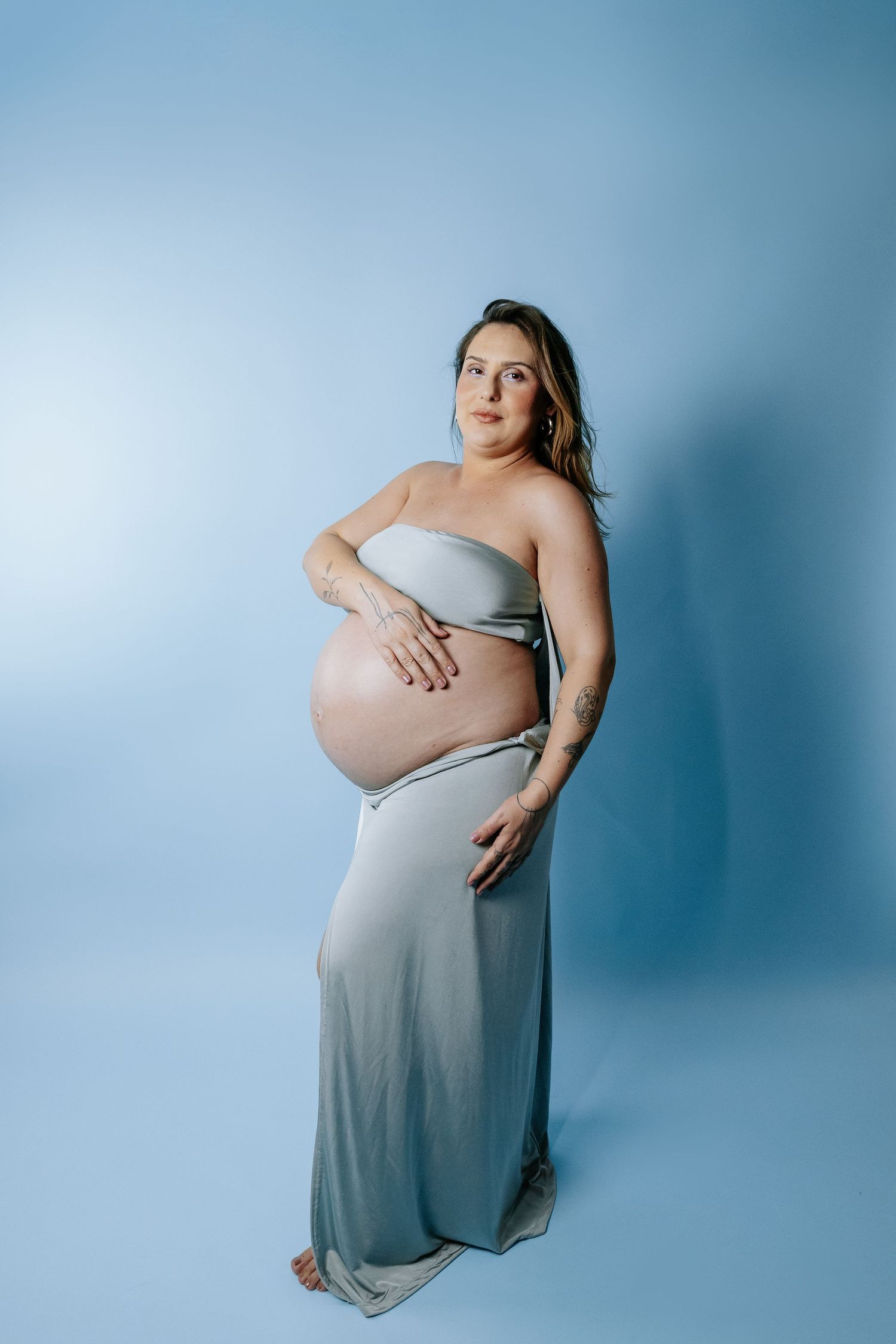 A maternity portrait featuring a grey flowing dress against a blue studio backdrop.