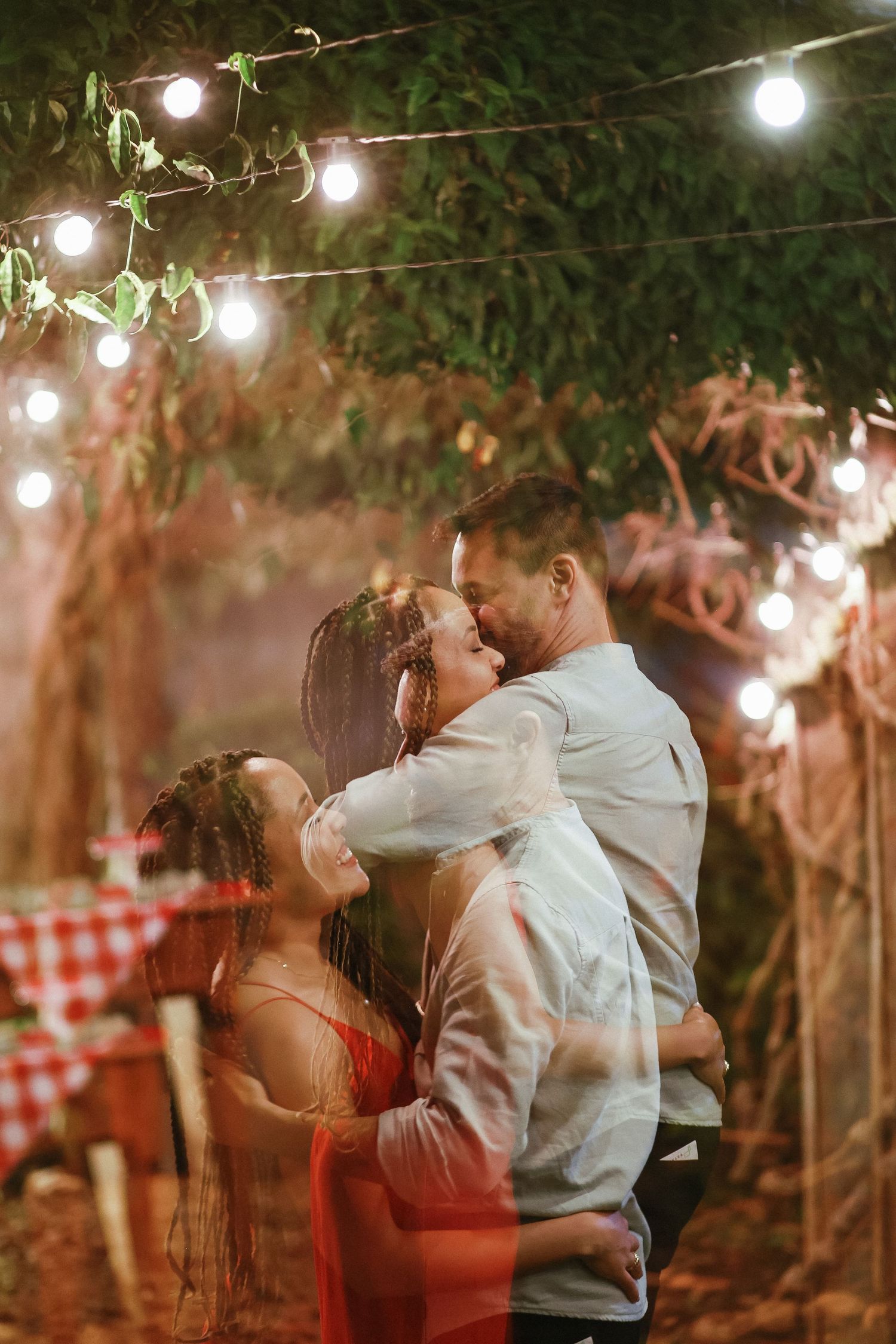 Couple embracing romantically under string lights in a lush garden setting at night, woman in red dress.