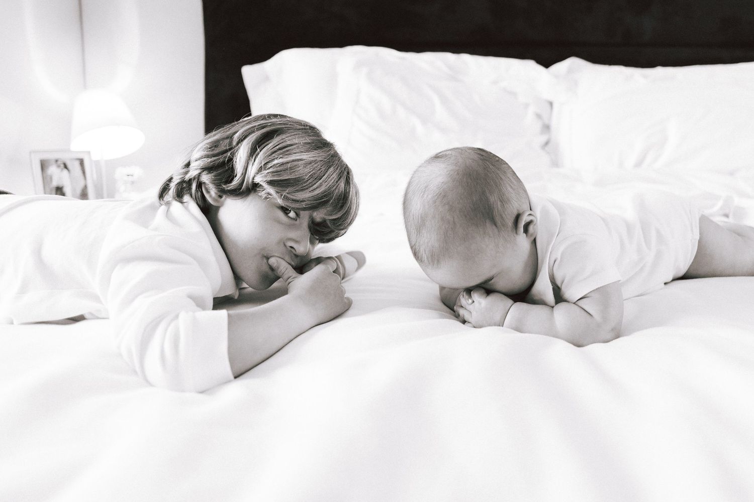 Black and white photo of a young child and baby lying on a white bed, facing each other in matching white outfits.