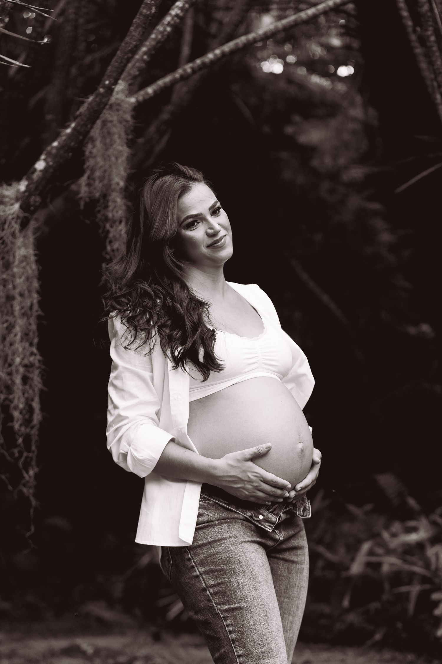 Black and white maternity photo of pregnant woman cradling baby bump outdoors in lush garden setting.