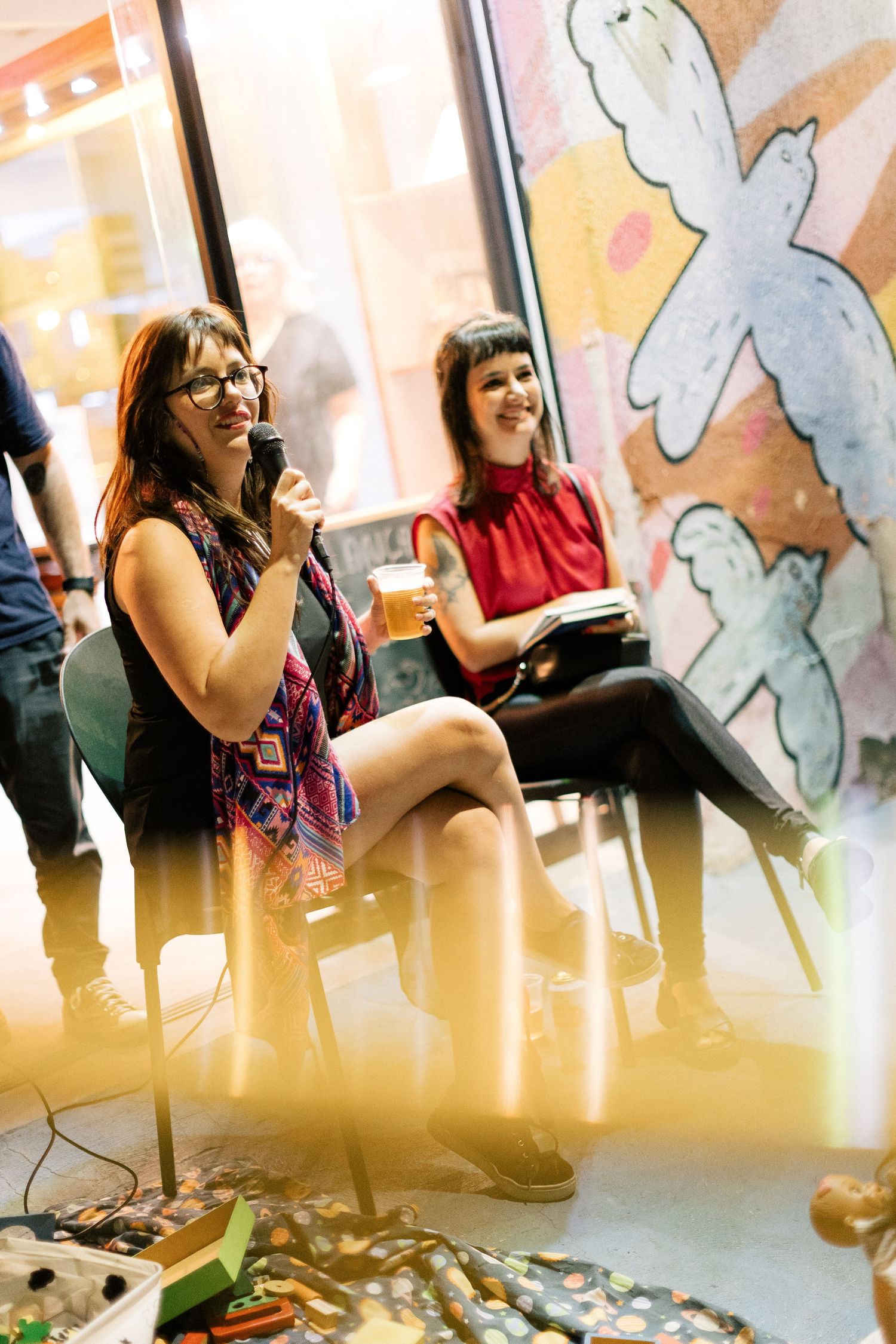 Two women laughing and socializing at an art gallery event, one holding a beer glass near colorful wall art.