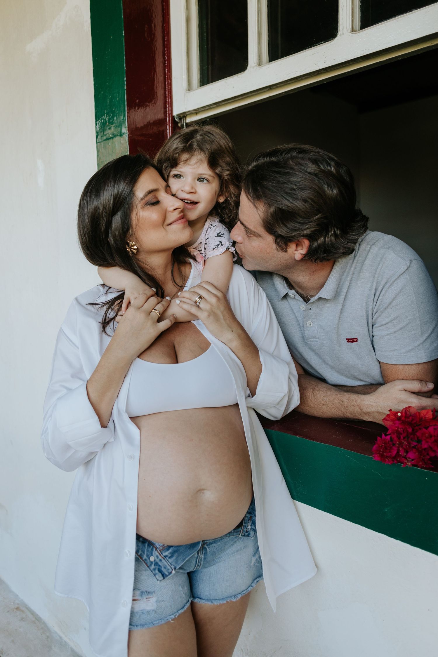 A couple shares romantic moments in front of a white wall and green trim in a multi-panel maternity photoshoot.