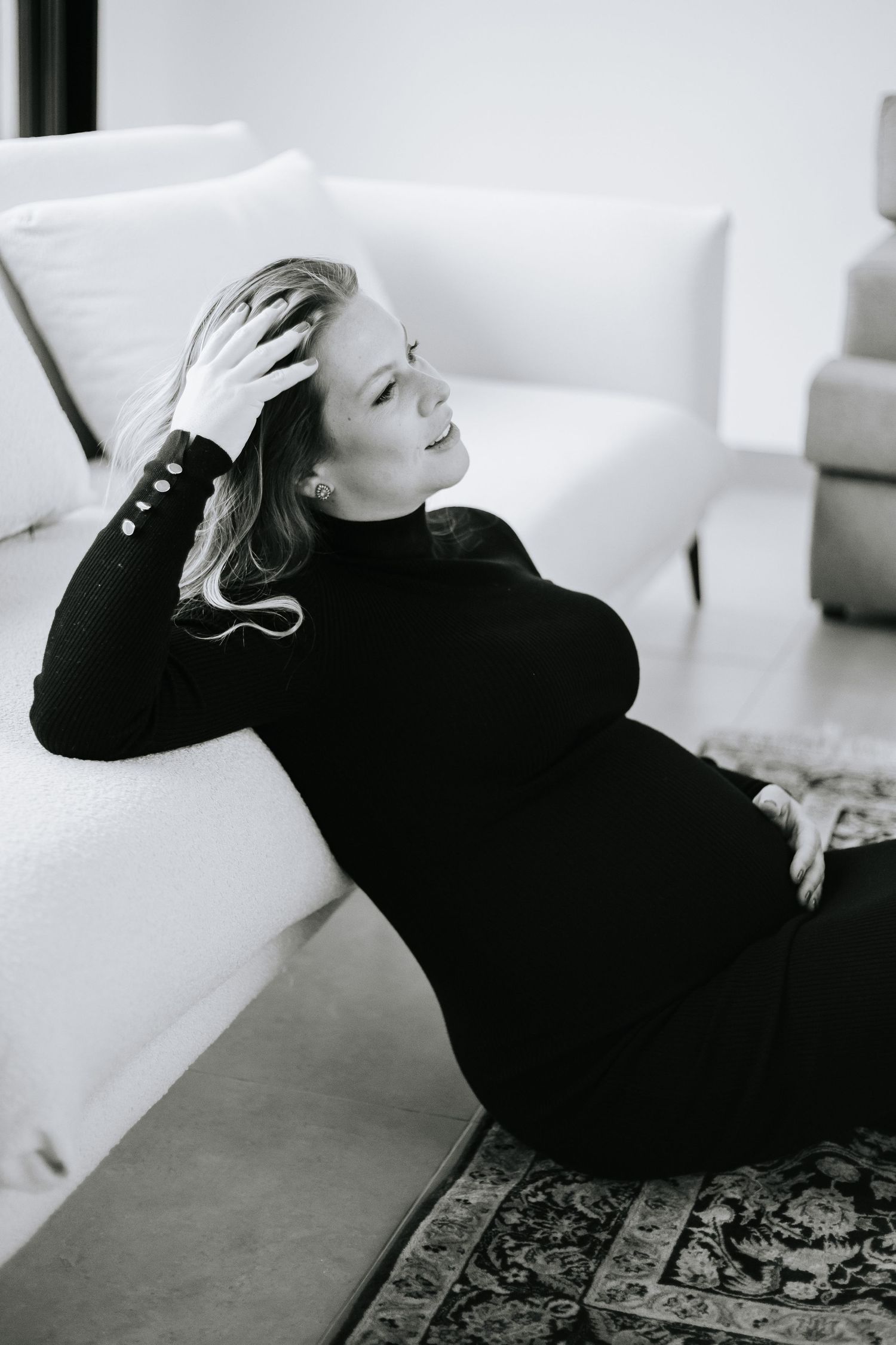 Black and white photo of pregnant woman in dark sweater reclining on white couch.