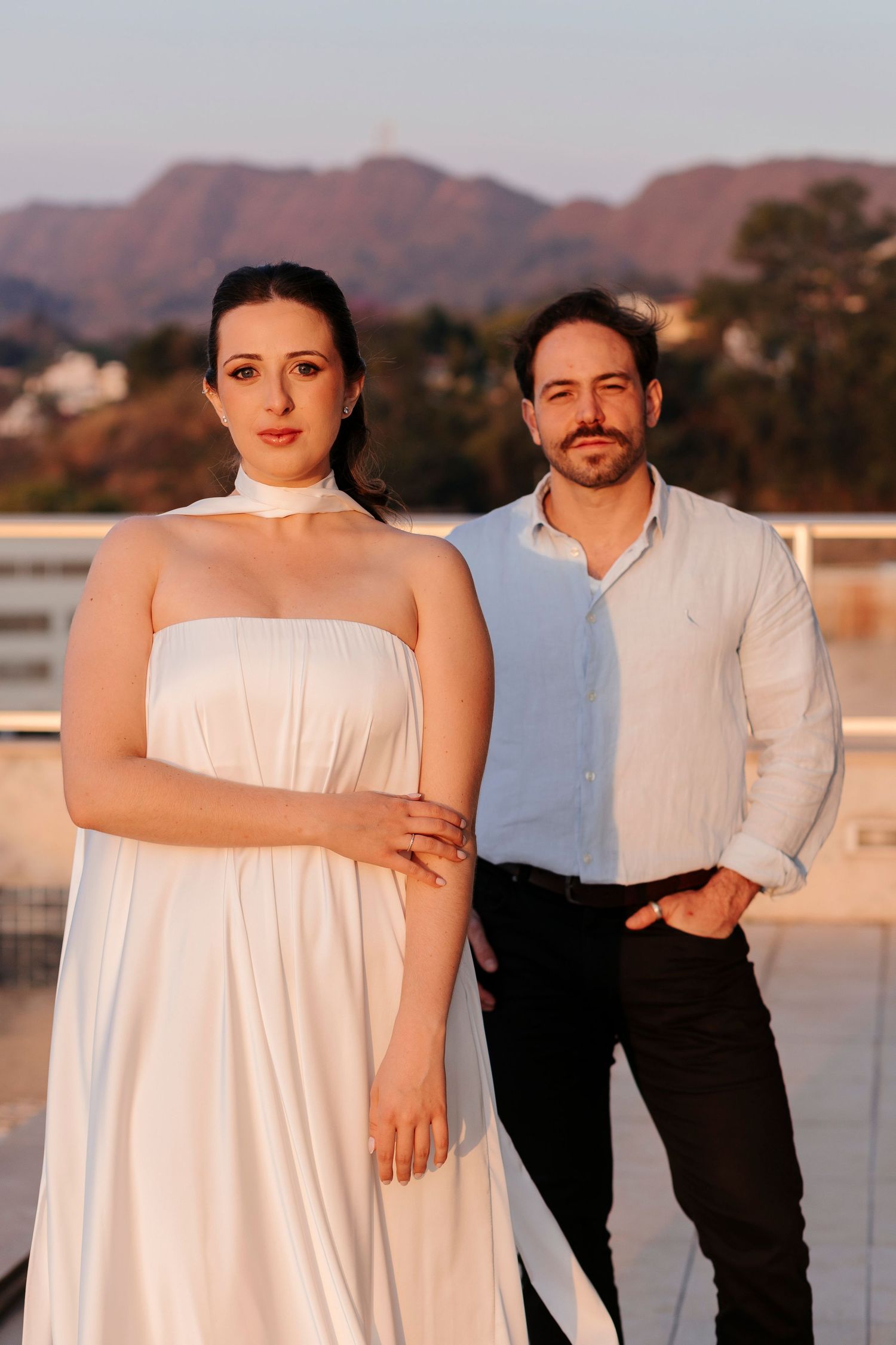Stylish couple in formal wear posed against mountain landscape at golden hour.