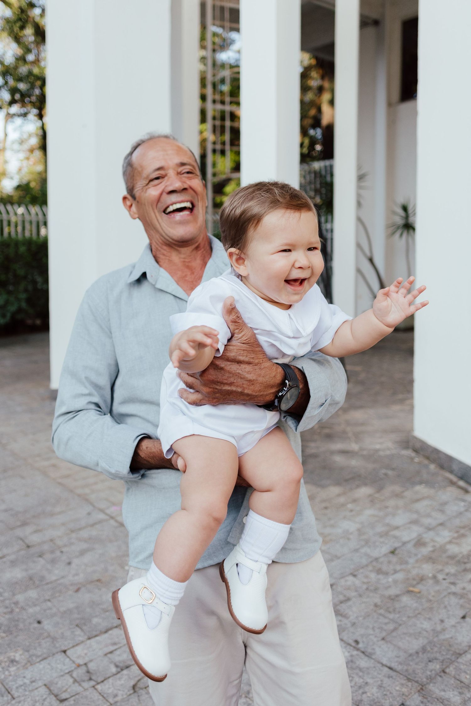 A joyful baby in white clothing shares laughter and playful moments with someone in a light blue shirt outdoors.
