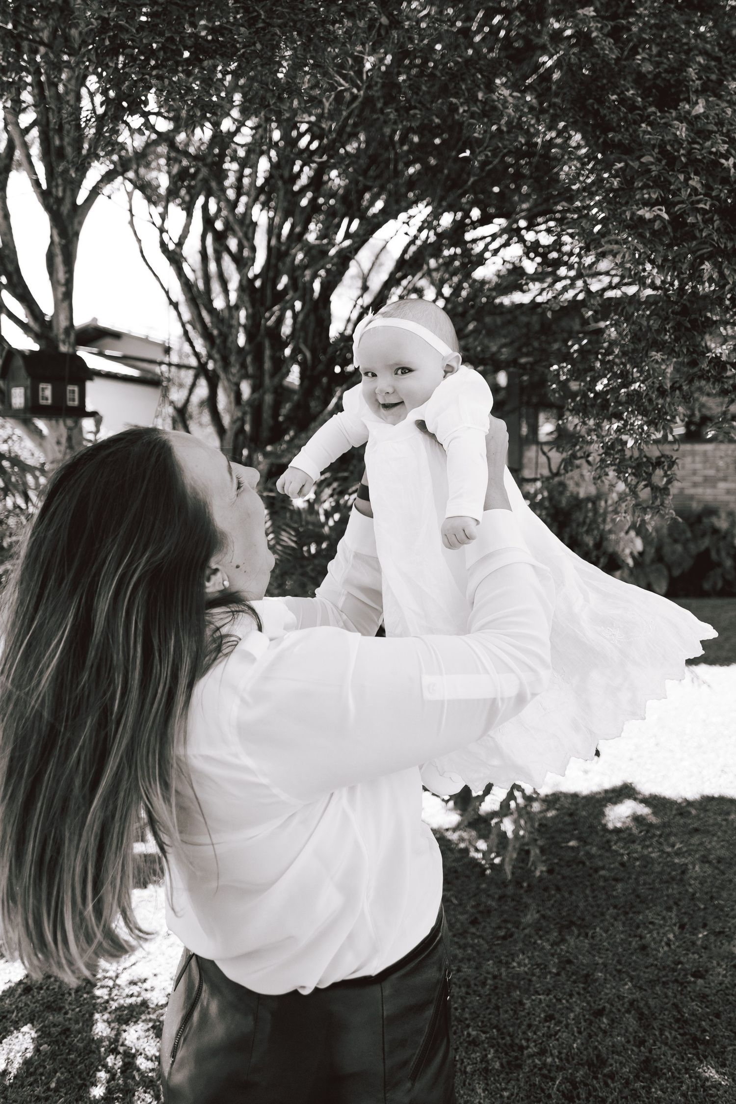 A black and white photo of a parent lifting a small baby in a white outfit outdoors in a garden setting.