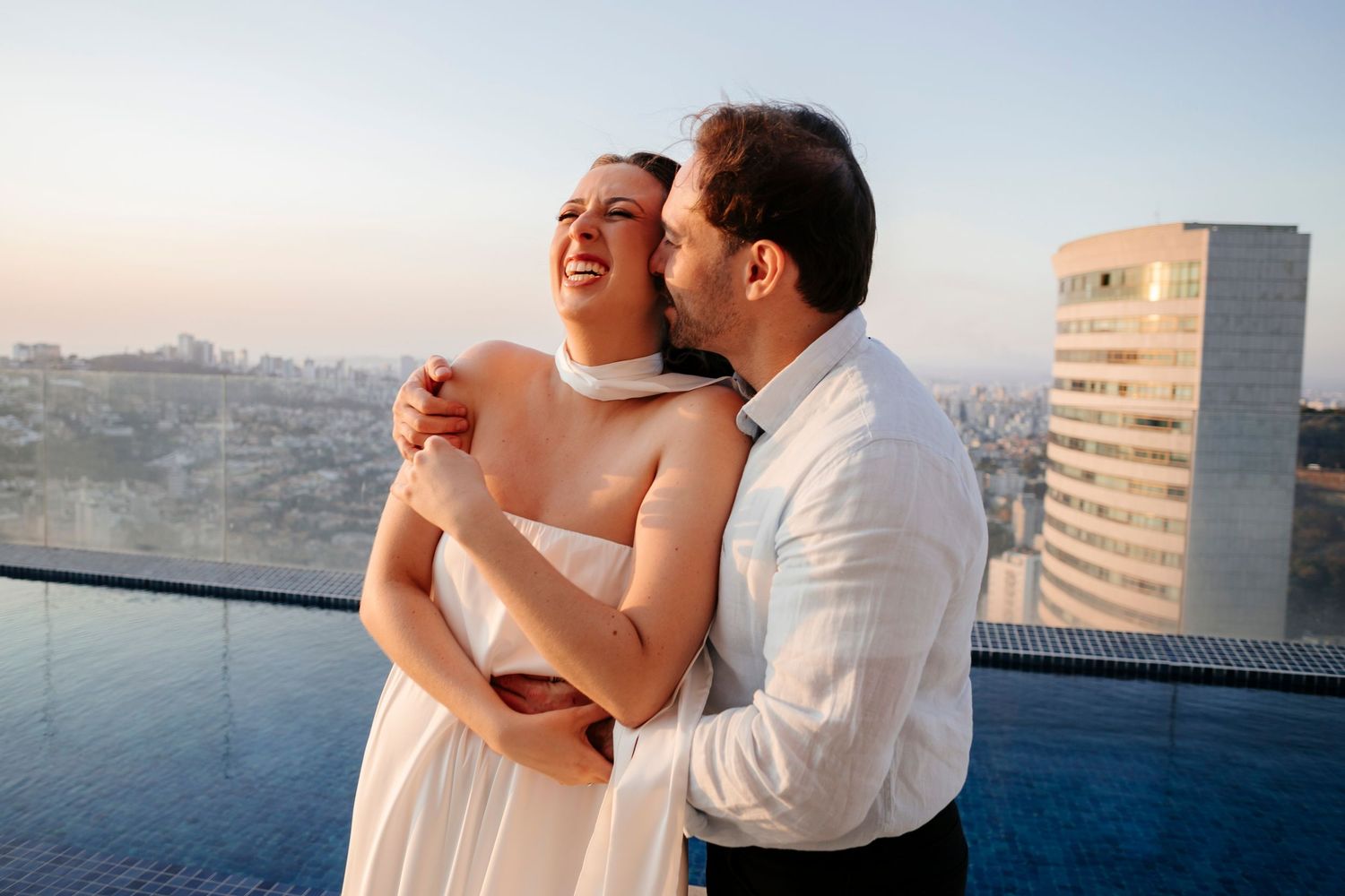 A couple shares romantic moments at sunset on a rooftop terrace overlooking a coastal cityscape.