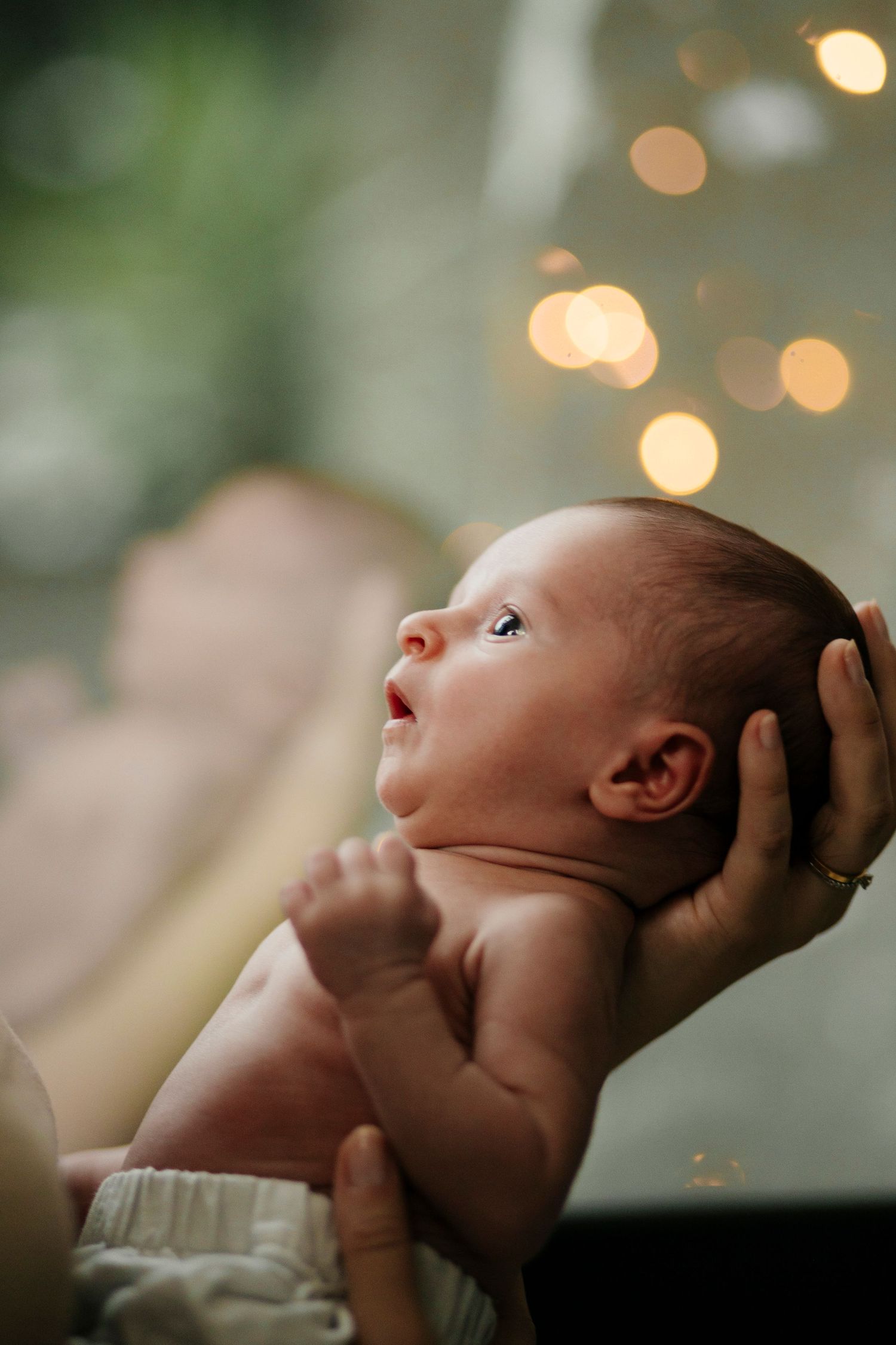 Newborn baby being held up against bokeh lights in artistic portrait.