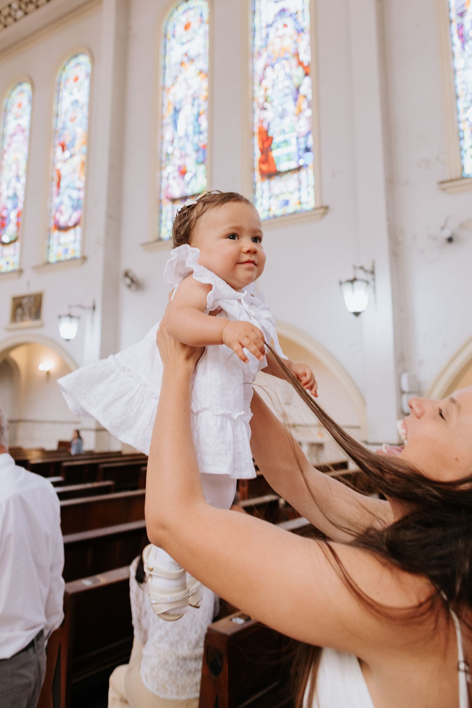 Baby in white outfit being lifted up inside a church with stained glass windows in the background.