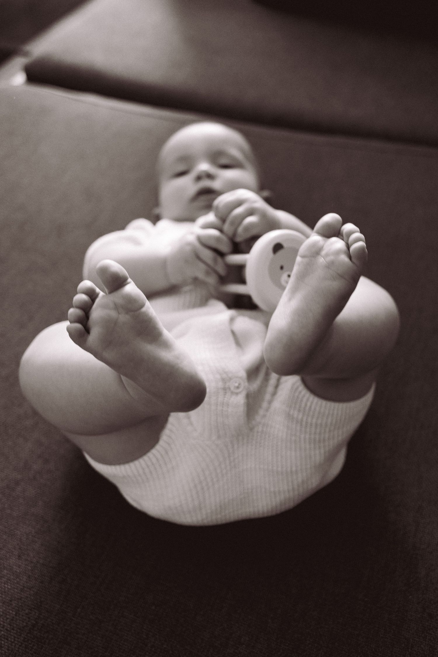 Black and white photo of a newborn baby lying on its back with tiny feet raised, holding a small toy.