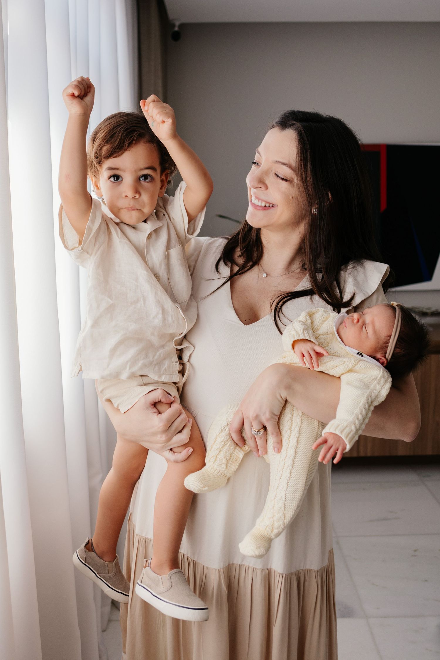 Smiling mother holding newborn baby and toddler son with raised arms by window.