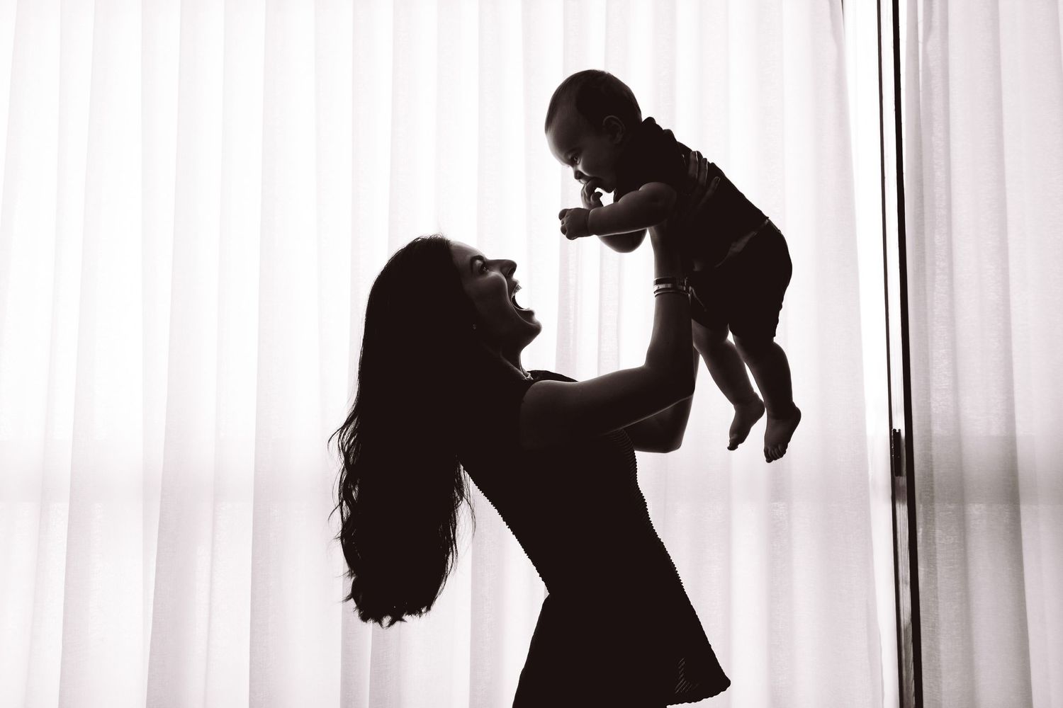 Silhouette of a mother lifting her baby in the air against a bright white curtain backdrop.