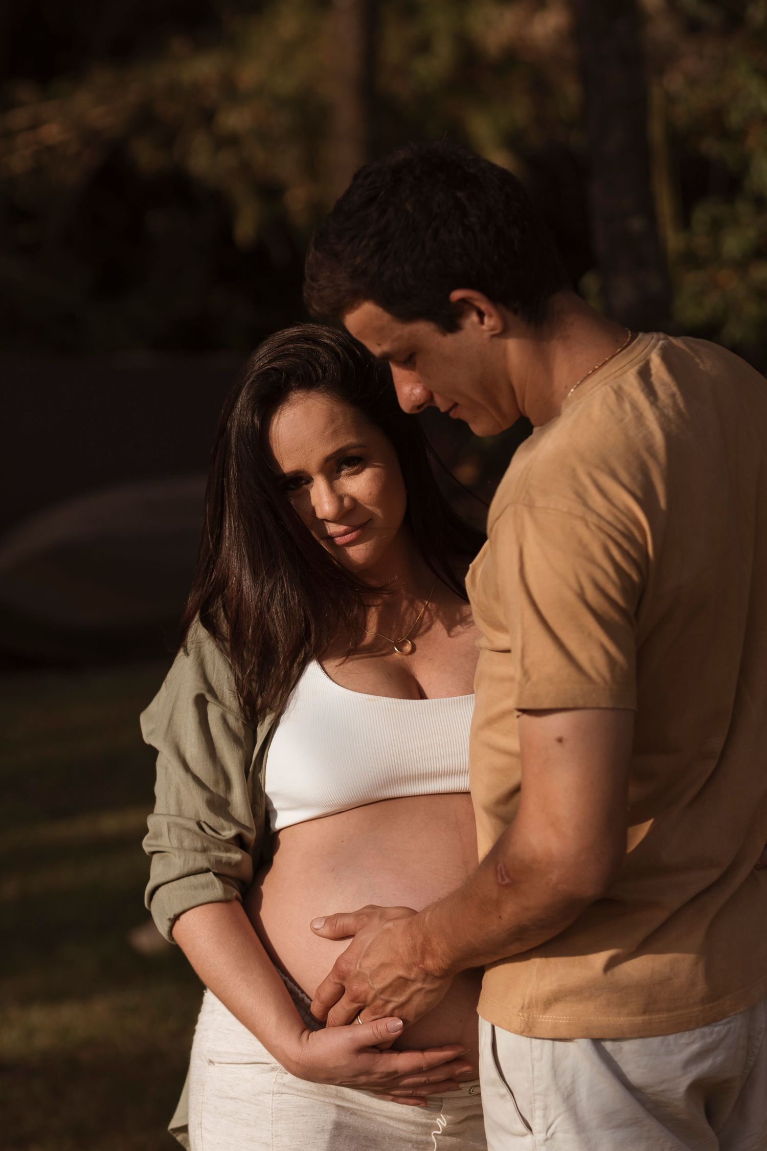 Intimate outdoor maternity photo of couple embracing and cradling baby bump at sunset.