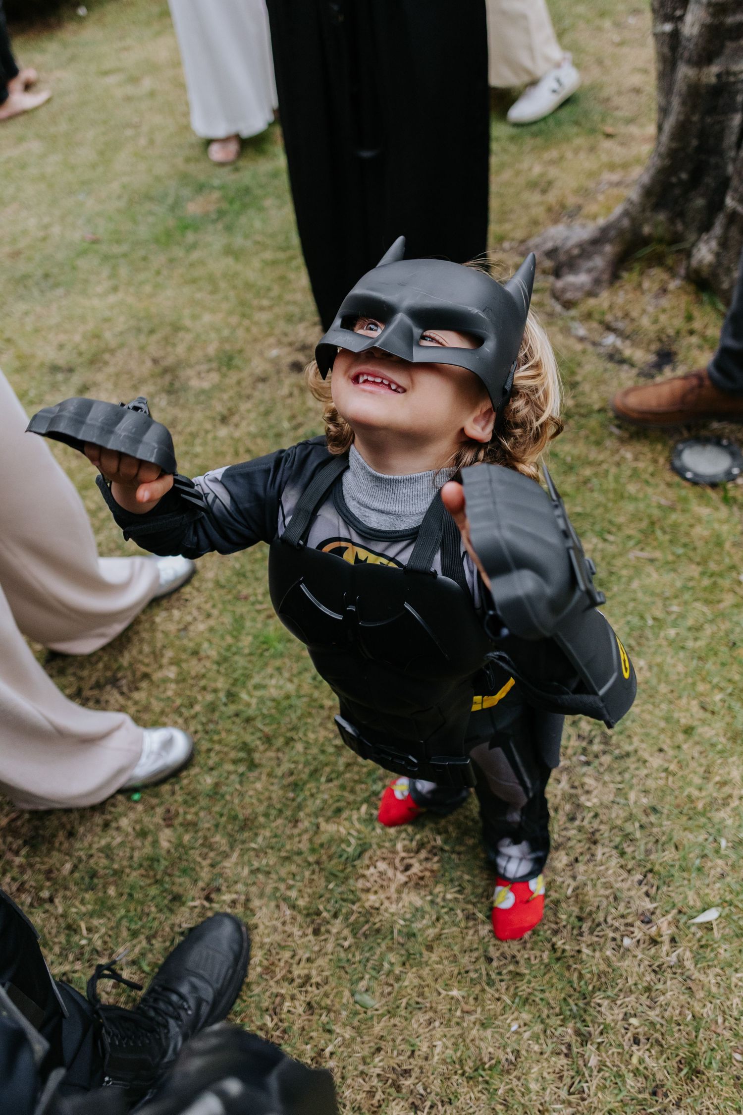 Person in Batman costume crouches on grass while drinking from a water bottle at an outdoor event.