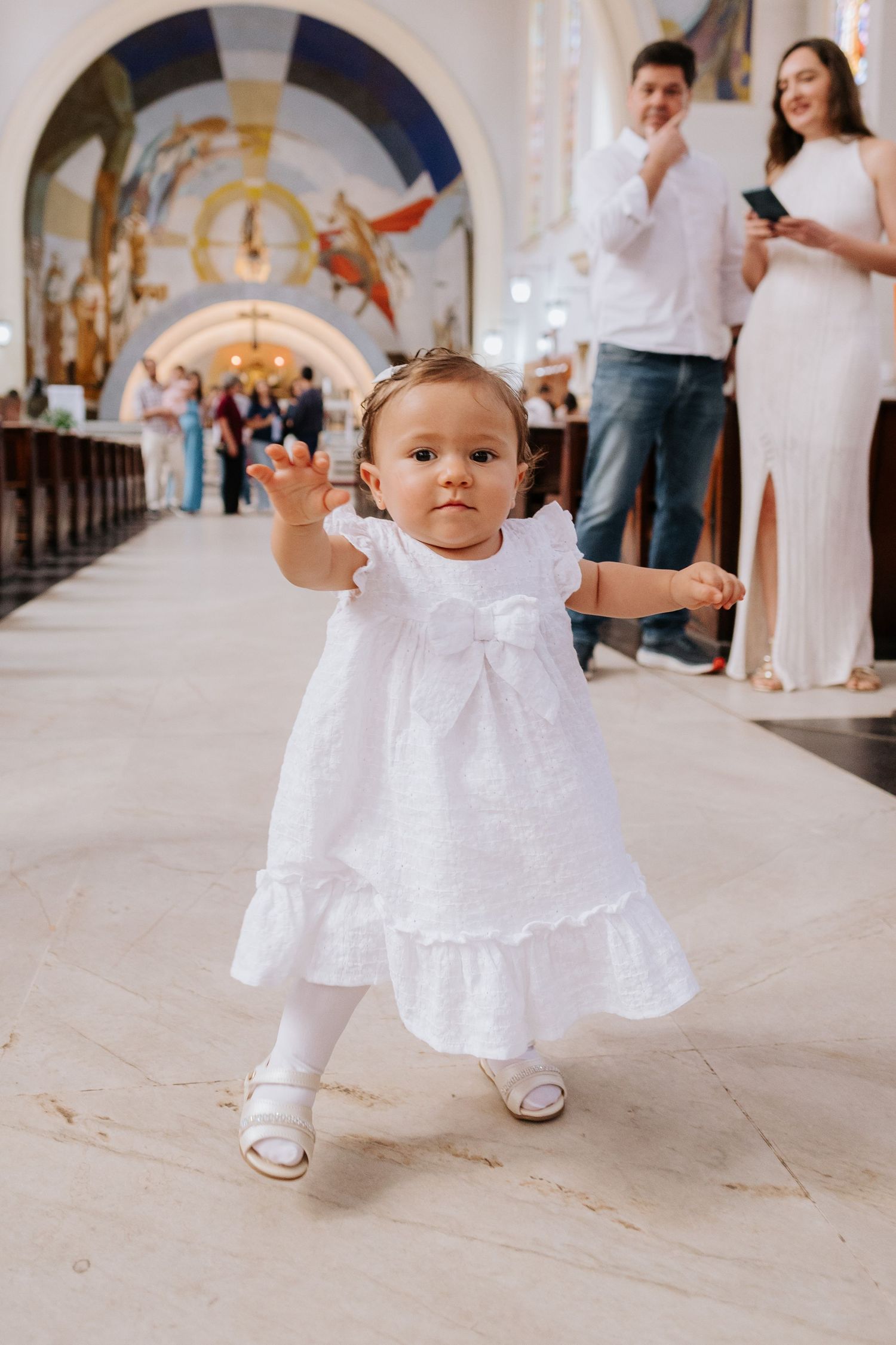 A toddler in a white baptismal dress takes first steps on a church floor with ornate arches in the background.