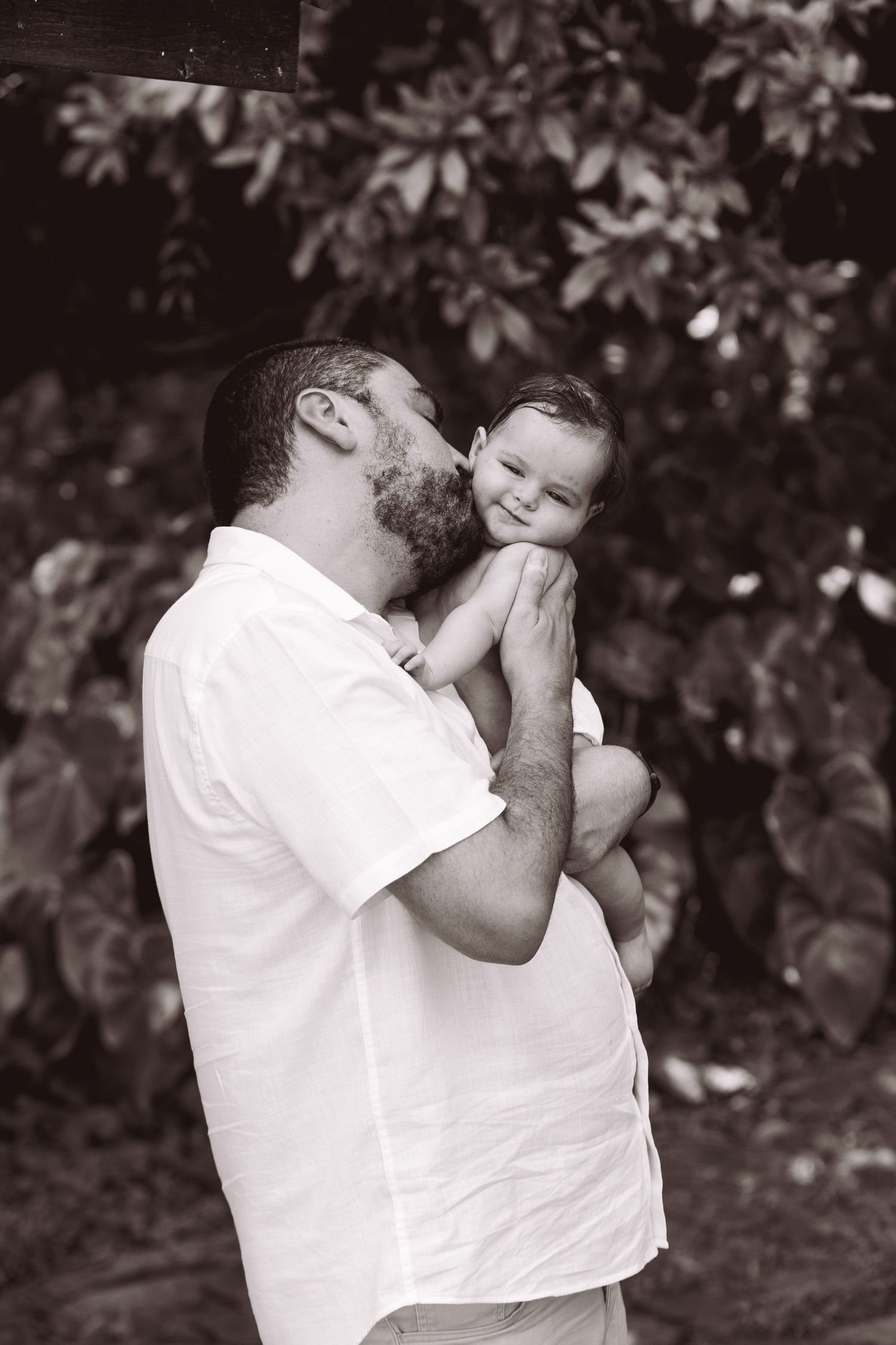 Black and white photo of a bearded father kissing his smiling baby while holding them outdoors near lush green foliage.