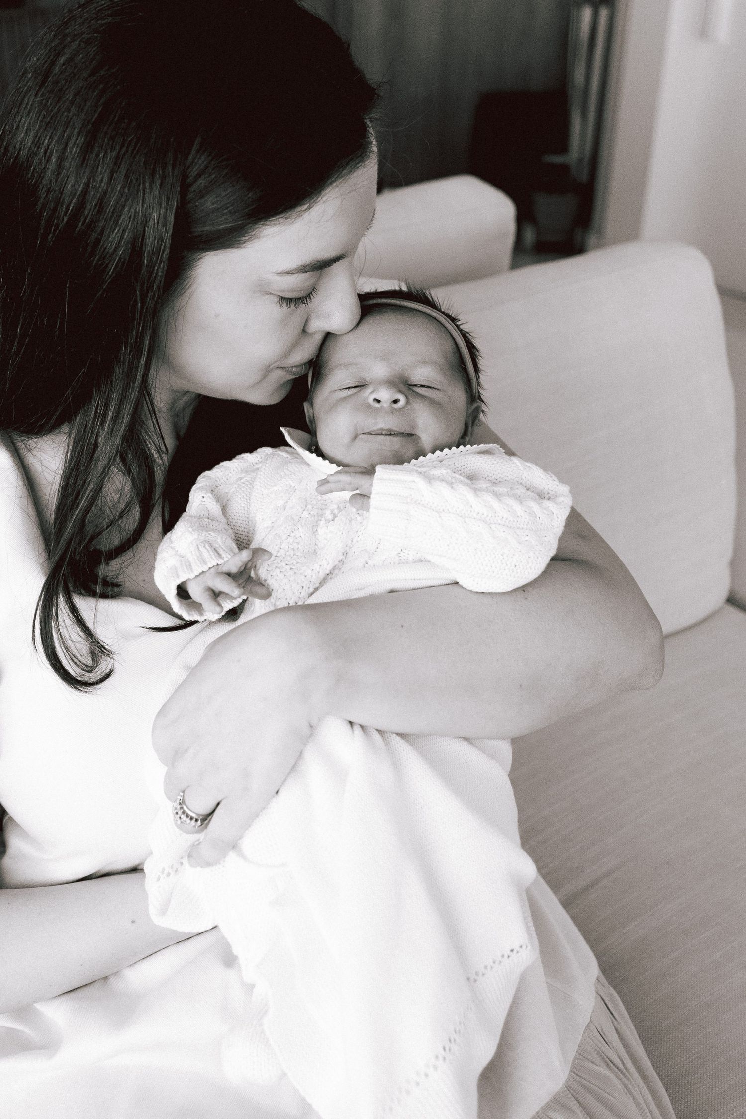 Black and white photo of a mother kissing her sleeping newborn baby wrapped in white.