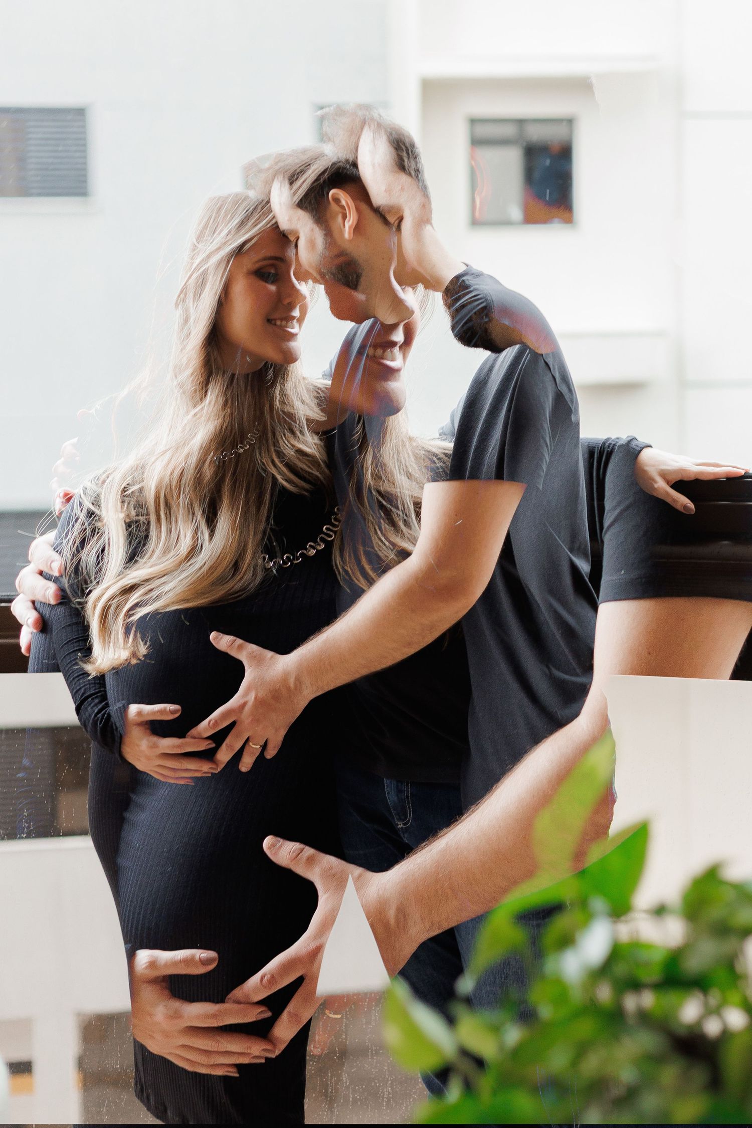 A happy couple in black outfits sharing a tender moment on a balcony, with the woman visibly pregnant.