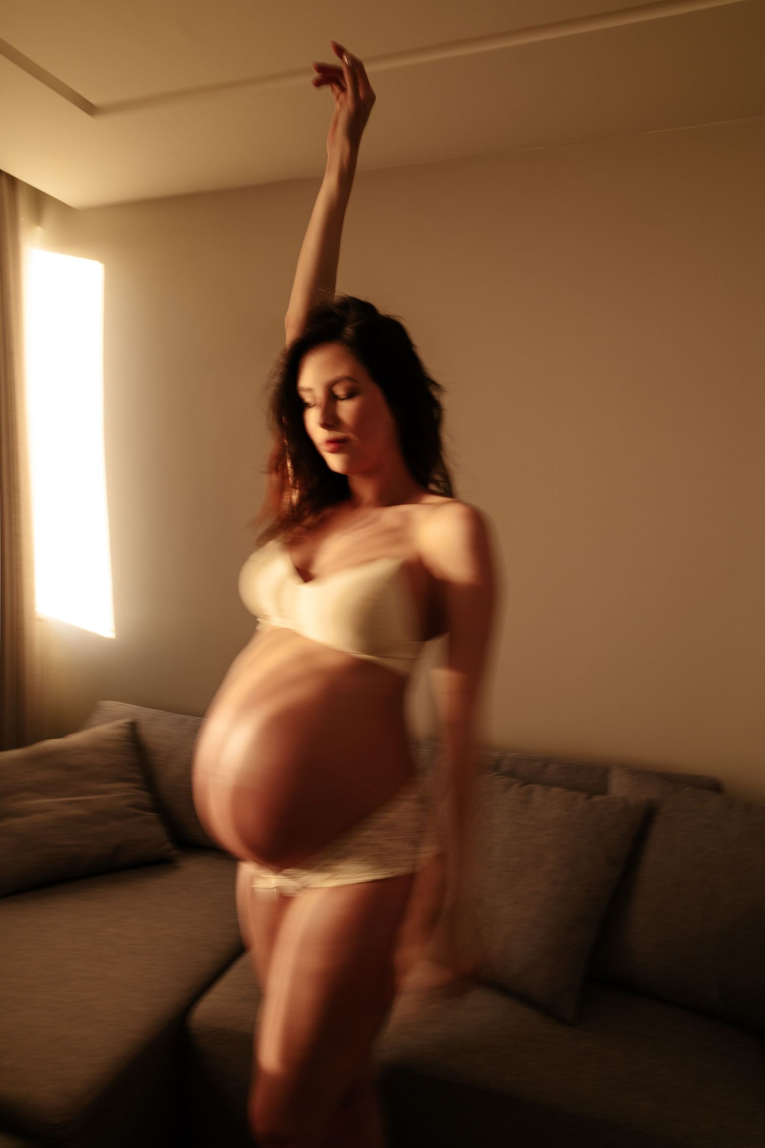 Graceful maternity pose with raised arm showing pregnant silhouette in warm lighting.