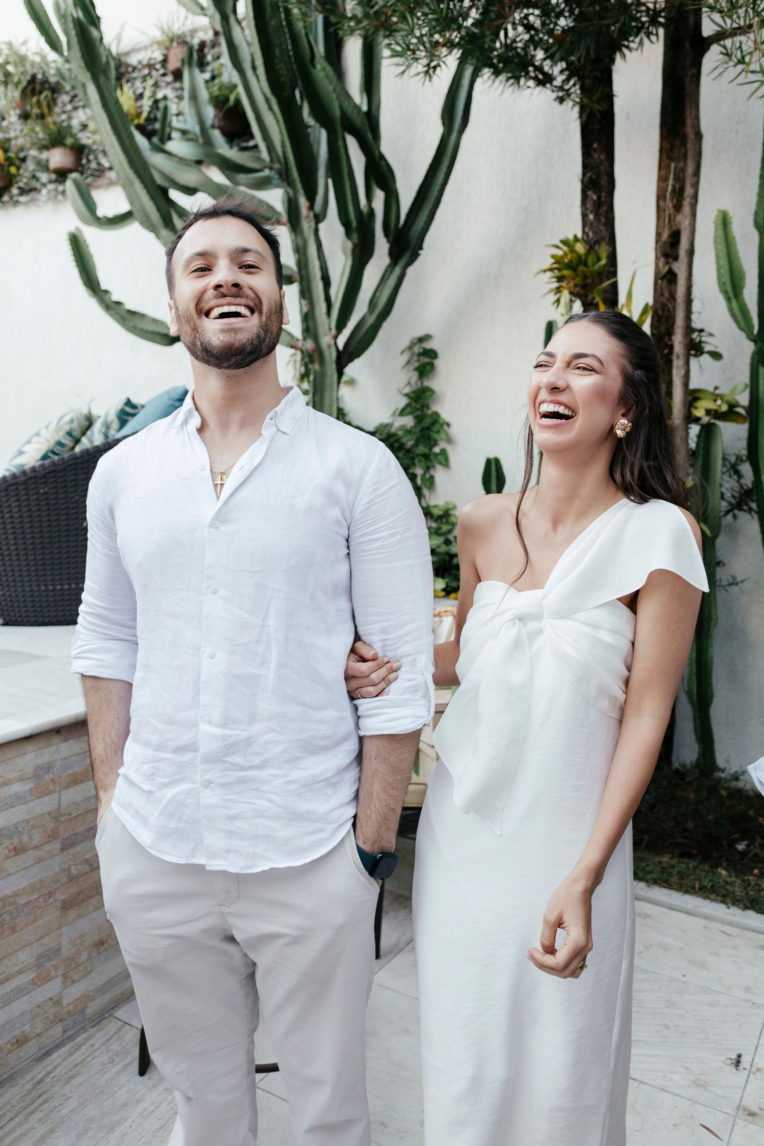 A couple shares a genuine laugh together against a white wall with cacti.