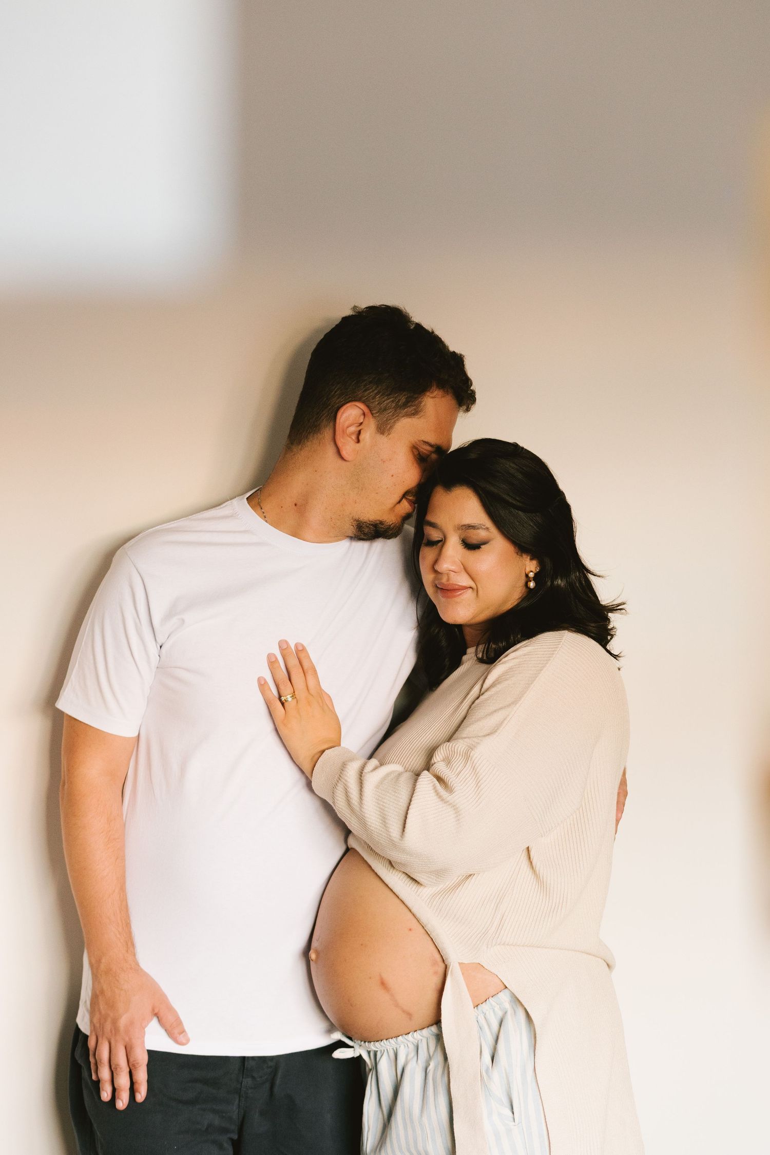 A couple sharing a tender moment, with a pregnant woman leaning into her partner against a soft neutral wall.