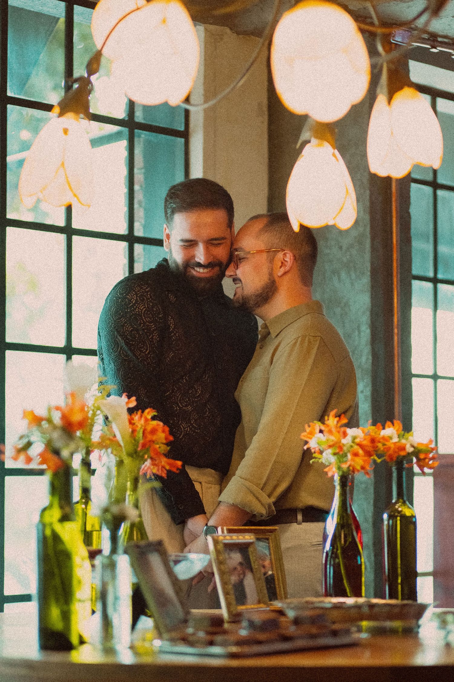 Two men share a tender moment in a warmly lit cafe with orange flowers and hanging lights.