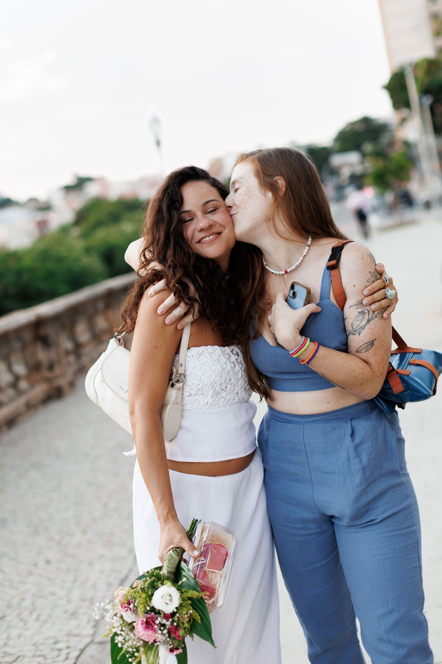 Two women hugging outdoors, one in white bridal outfit holding flowers, other in blue crop top.