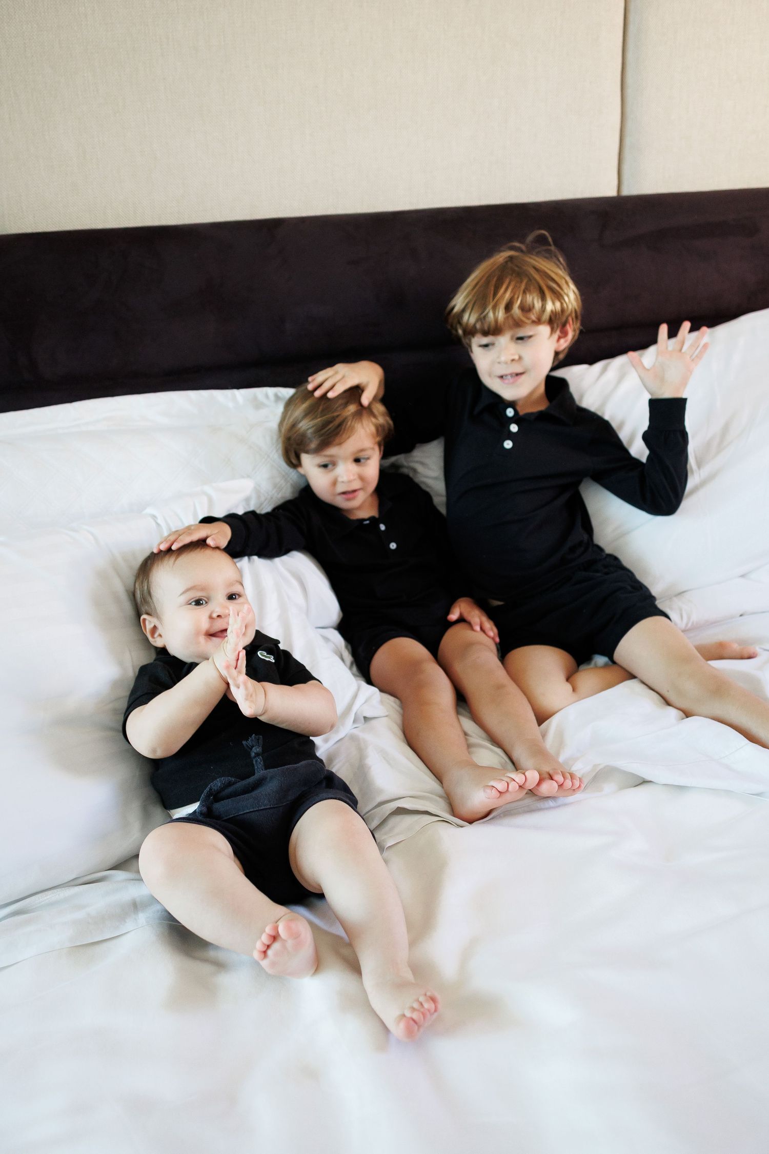 Three young boys in black and white outfits sitting together on a white hotel bed, smiling.
