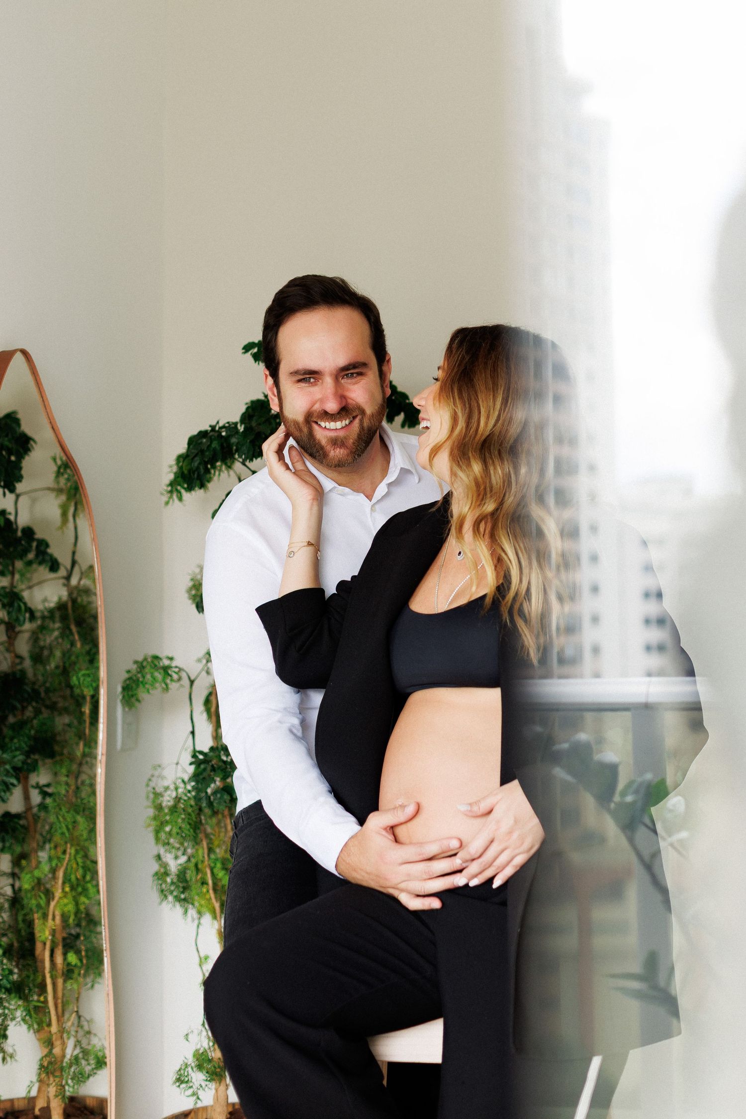 A smiling man in a white shirt embraces a pregnant woman in black near a window with green plants in the background.