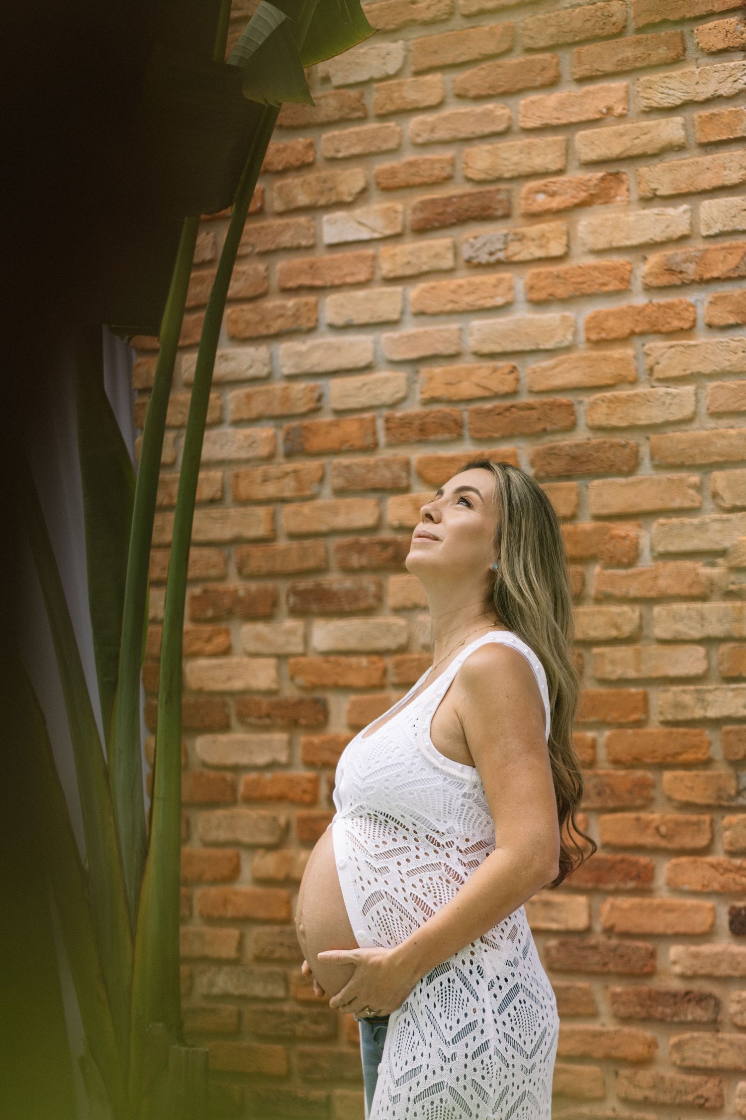 A person in a white lace dress poses against a rustic brick wall with dramatic lighting and a plant accent.