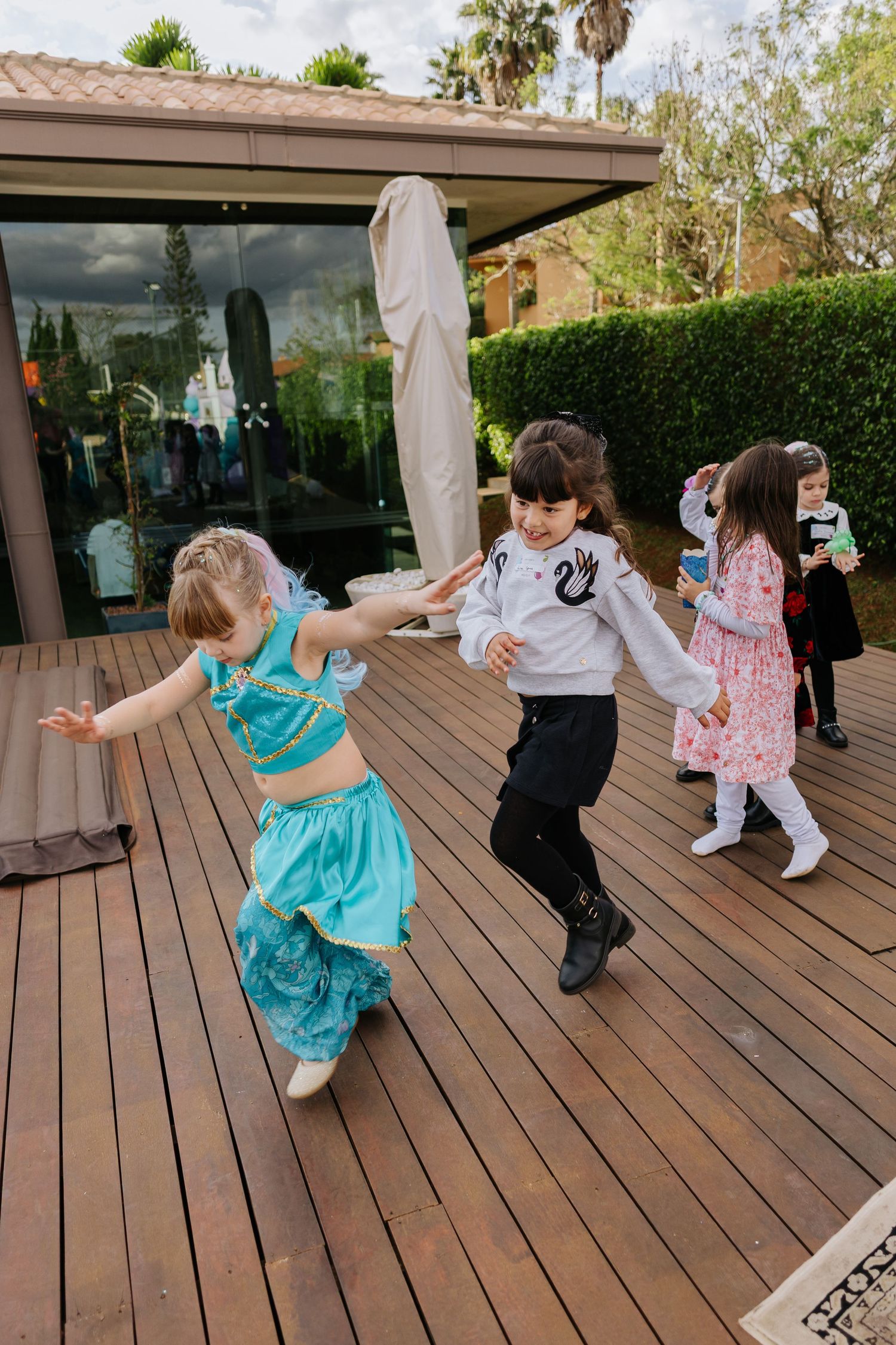 Children dancing together on a wooden deck at an outdoor party with palm trees in the background.