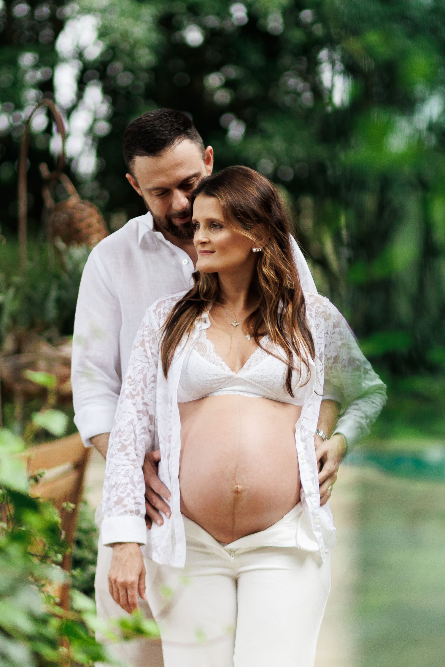 A couple in white outfits poses outdoors, the man embracing a pregnant woman showing her baby bump in a lush green garden.