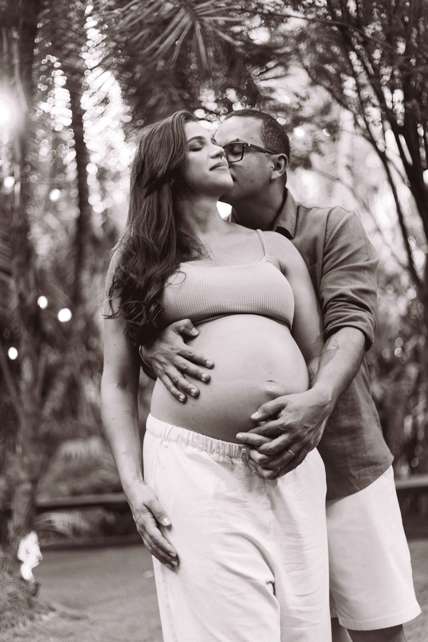 Black and white photo of a man kissing a pregnant woman while cradling her baby bump outdoors.